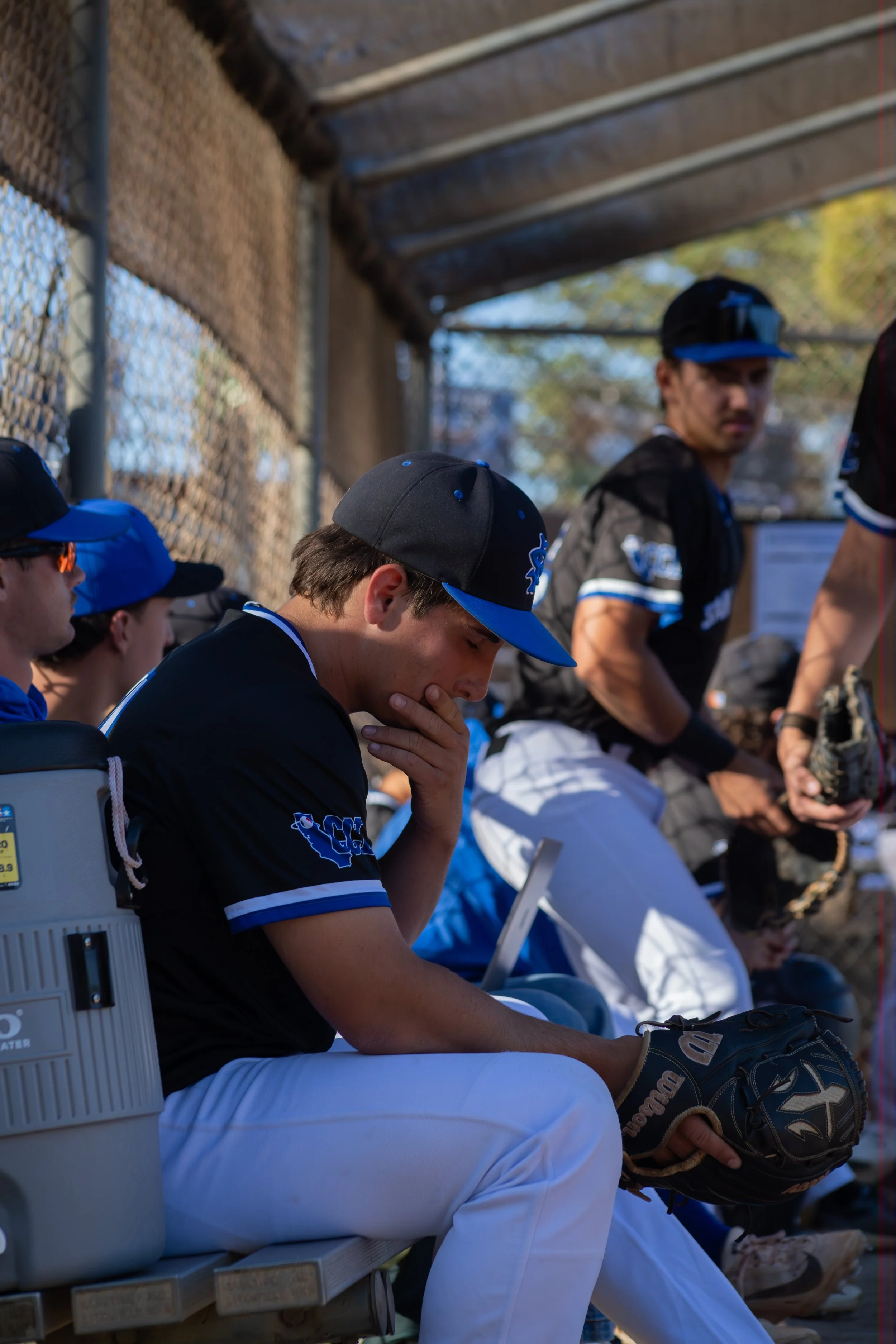 A group of young baseball players sitting on a bench, wearing team uniforms and caps, with one player in the foreground looking contemplative. The background shows a chain-link fence and some trees.
