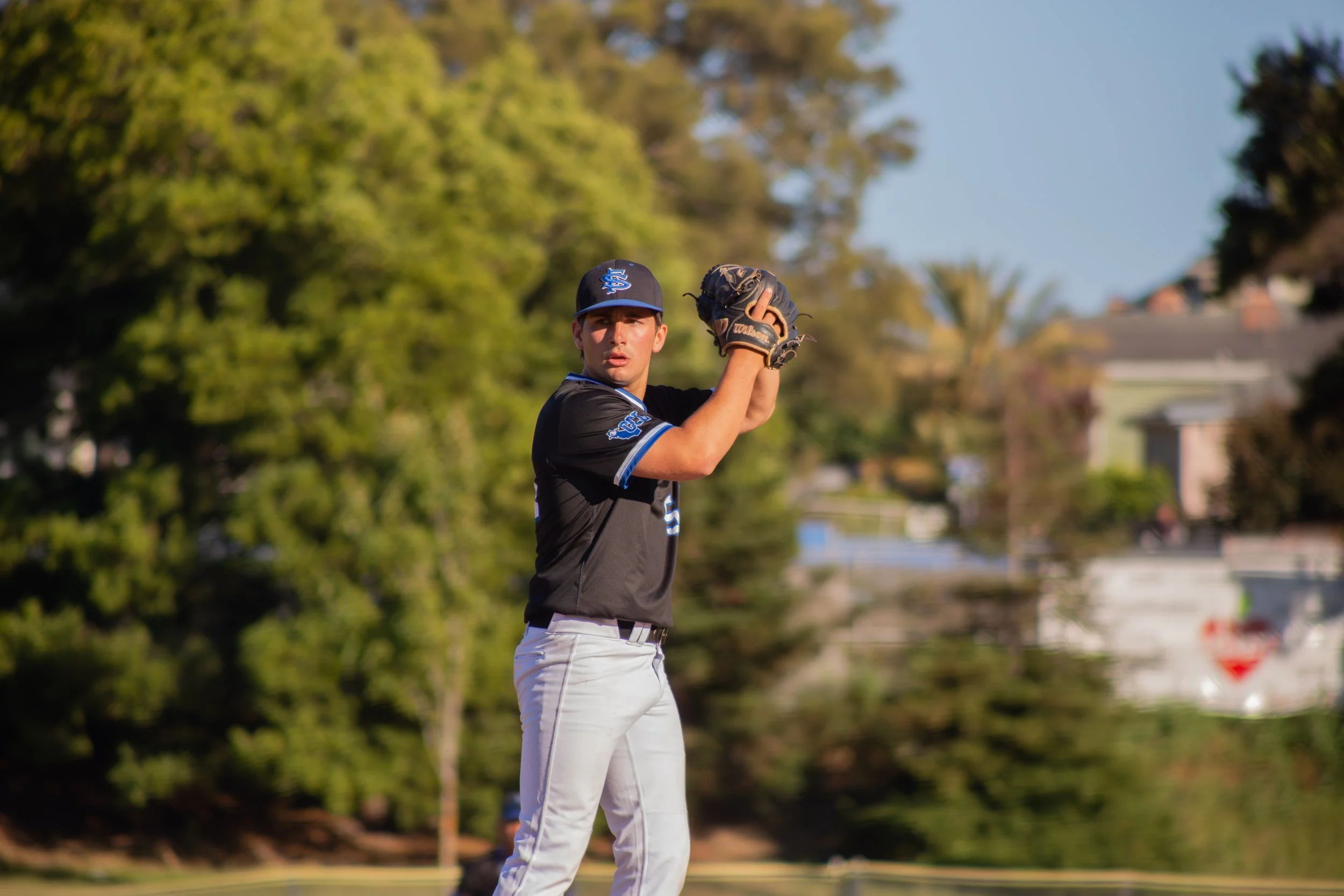 Young male baseball player in uniform with cap and gloves preparing to catch a ball outdoors on a grassy field with trees and houses in the background.