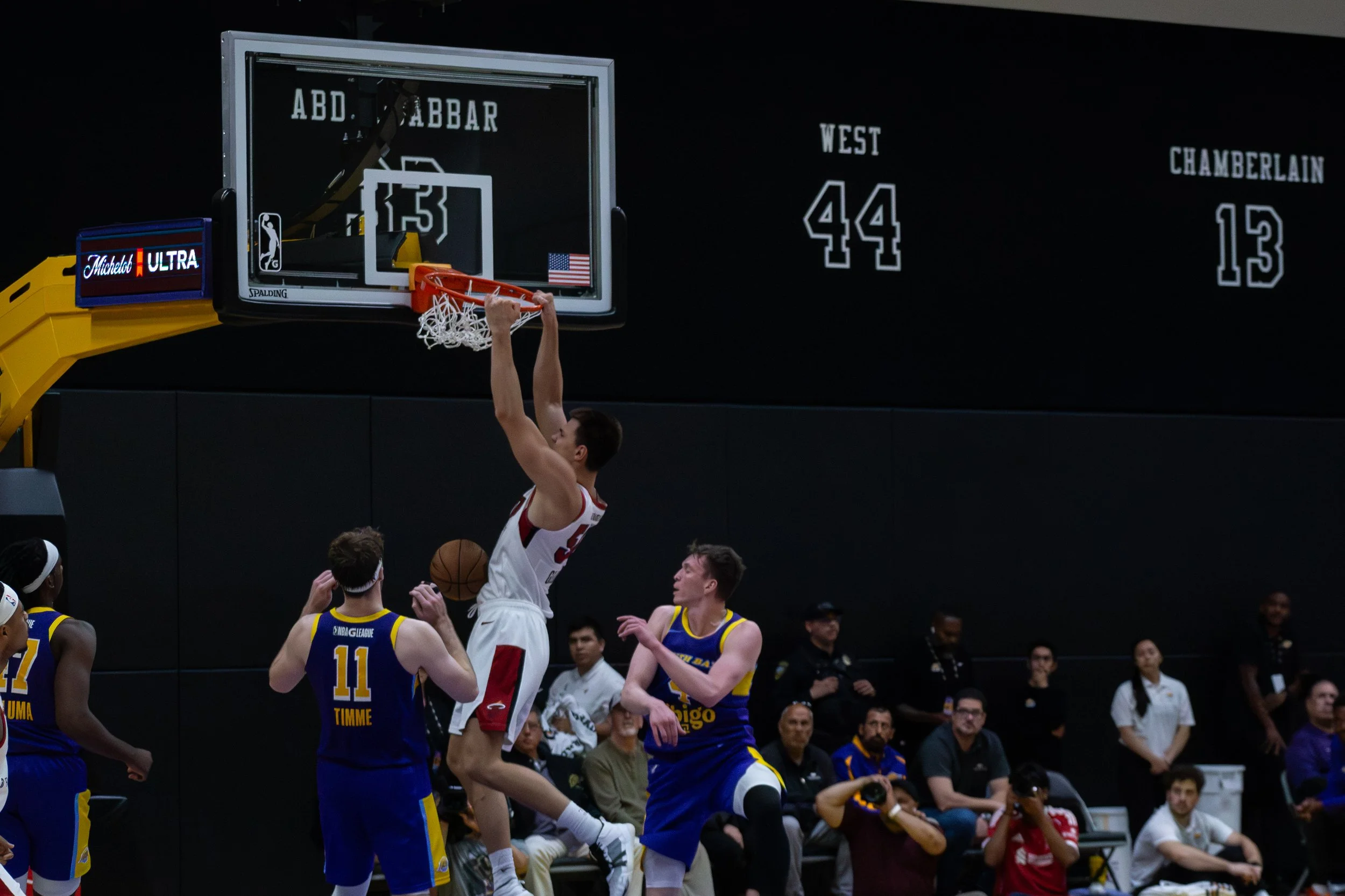 A basketball game with players competing near the basket, some players are wearing yellow and blue jerseys while one is in a white and red jersey attempting a shot, and others are watching or defending.