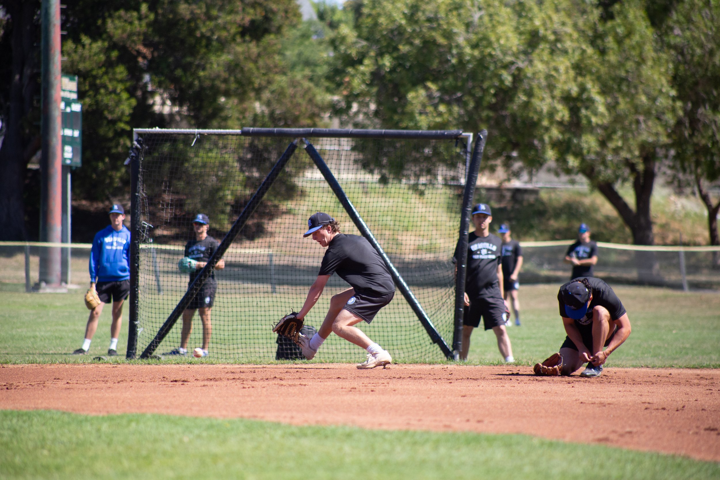 Young baseball players practicing on a field, some in baseball gear, with a focus on a player near the home plate in a black uniform. Spectators or team members watch in the background.