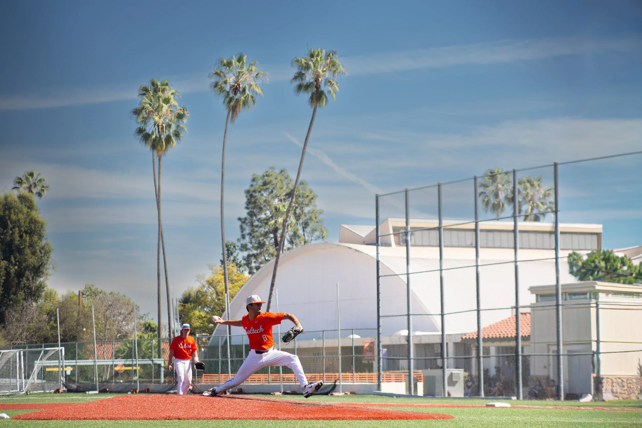 A baseball player in a red uniform pitches on the field as another player in red prepares to catch in the background, with blue sky, palm trees, and a baseball field surrounding them.