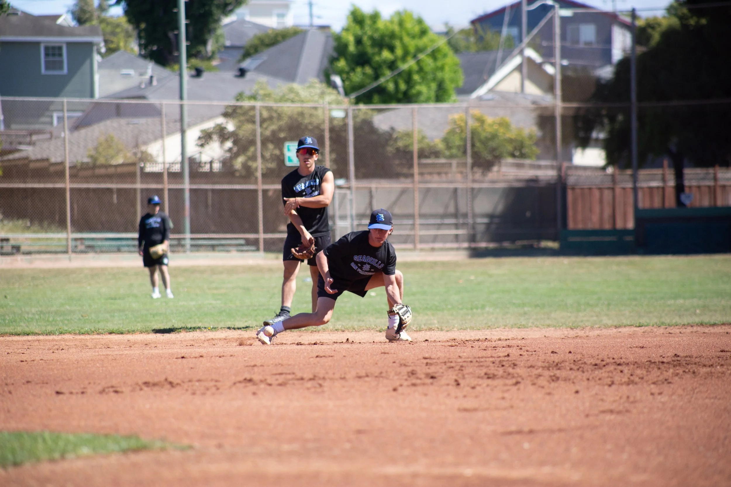 A baseball player in a black uniform and cap is diving to catch a ball on a baseball field with a dirt infield and green grass. In the background, another player stands near a chain-link fence with residential houses and trees beyond.