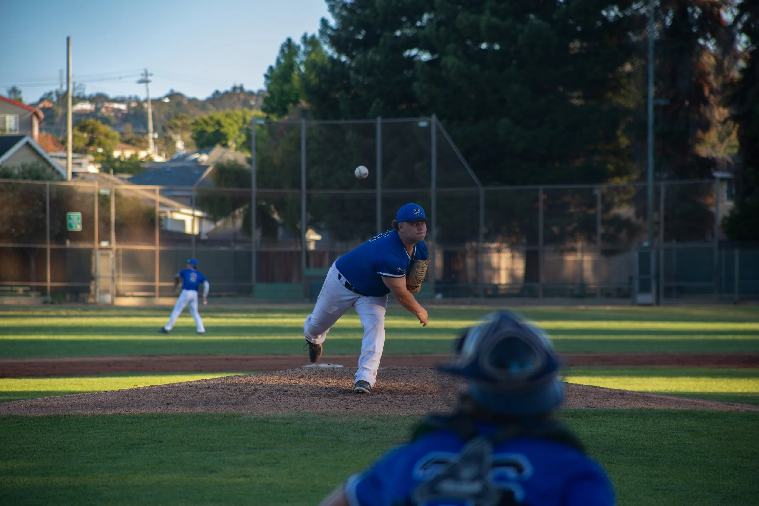 A baseball player in a blue uniform is pitching on a baseball field with a ball in mid-air. Another player is in the background, and a camera lens is in the foreground. The scene is outdoors with trees and houses in the distance, and it appears to be