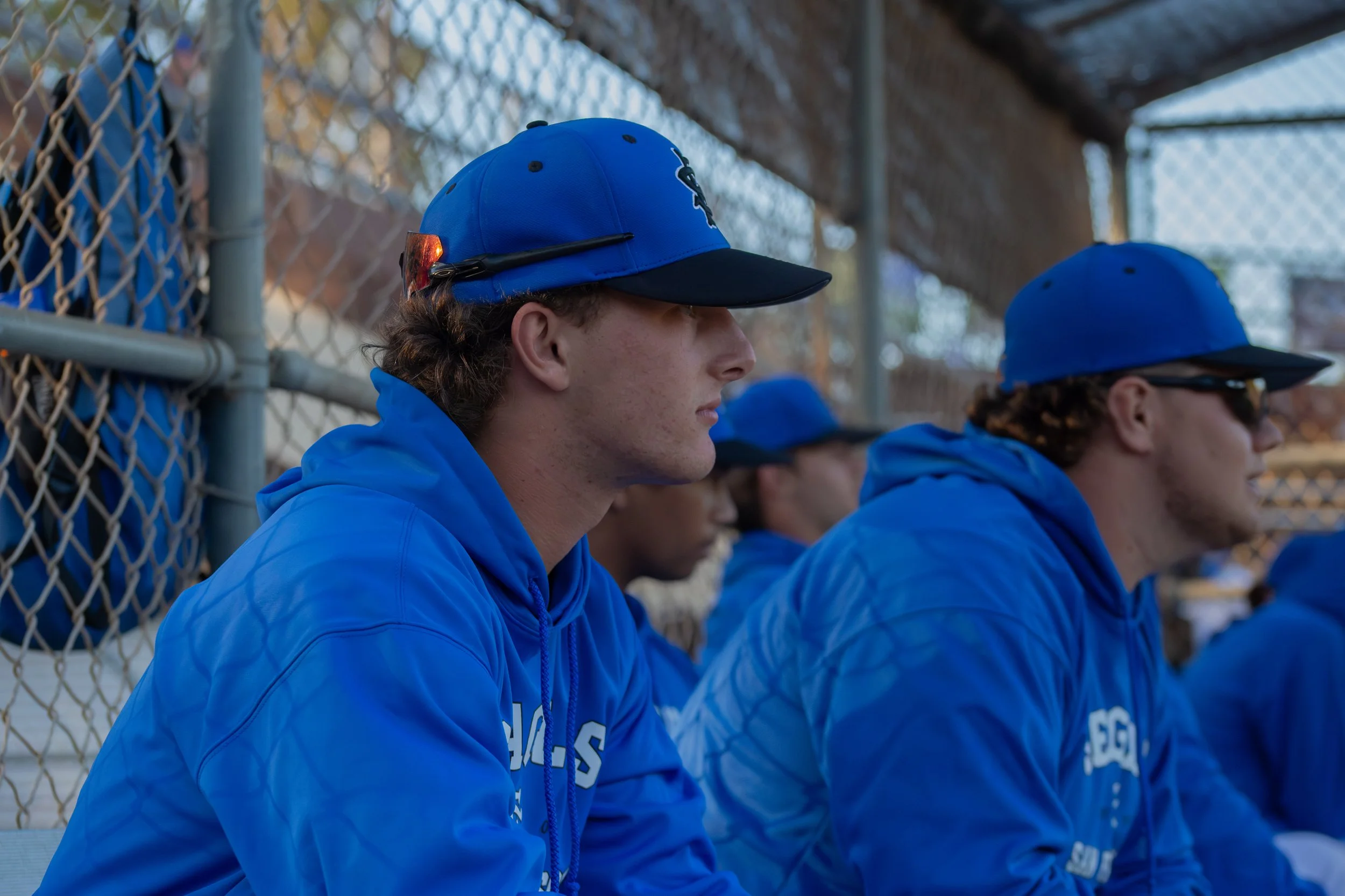 A group of young male baseball players wearing blue uniforms and caps sitting on a bench in a dugout, focused and attentively watching the game.