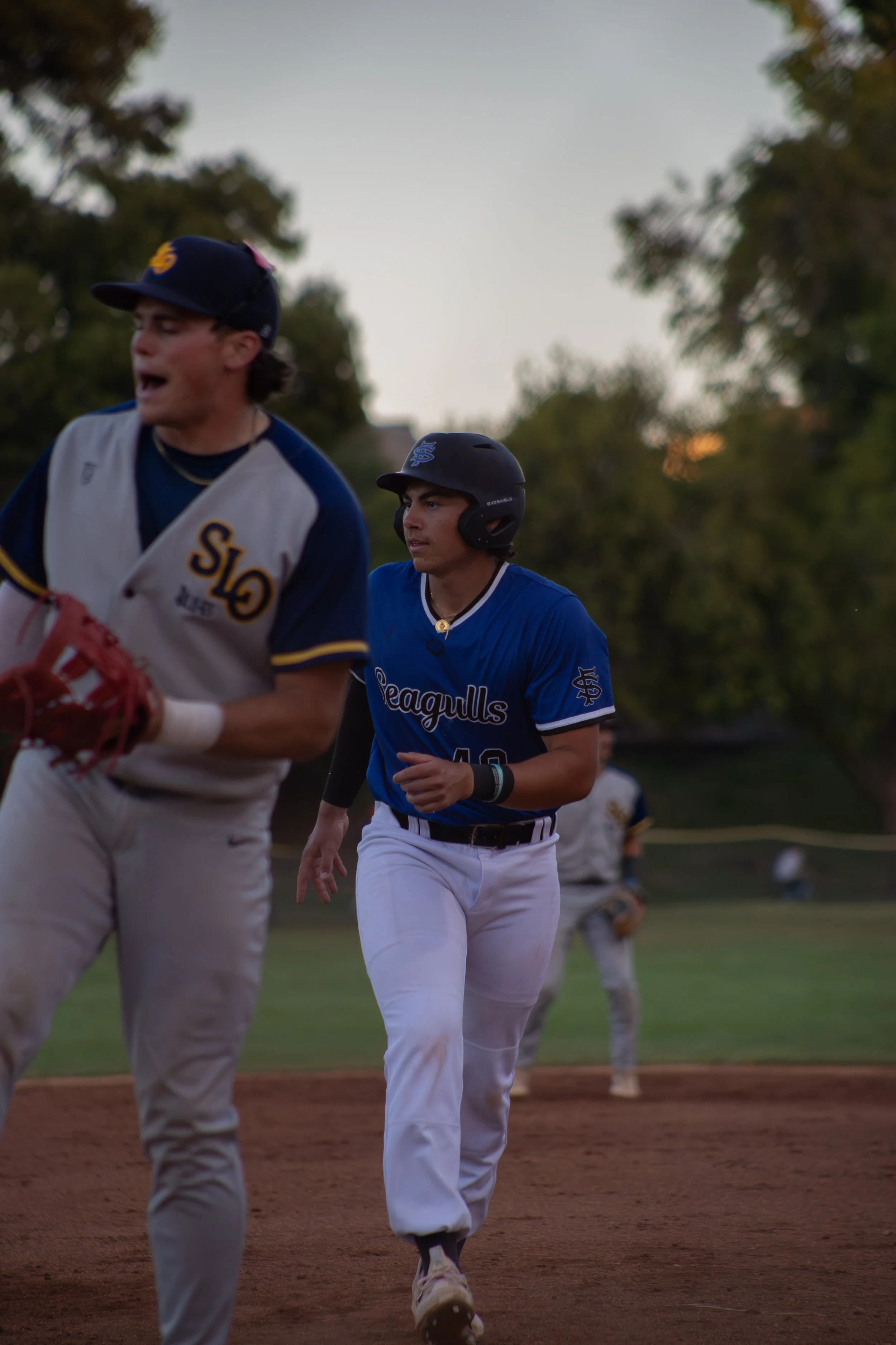 Two baseball players on the field during a game, with a grassy area and trees in the background, one in a blue uniform and the other in a cream uniform, both wearing helmets and white pants.