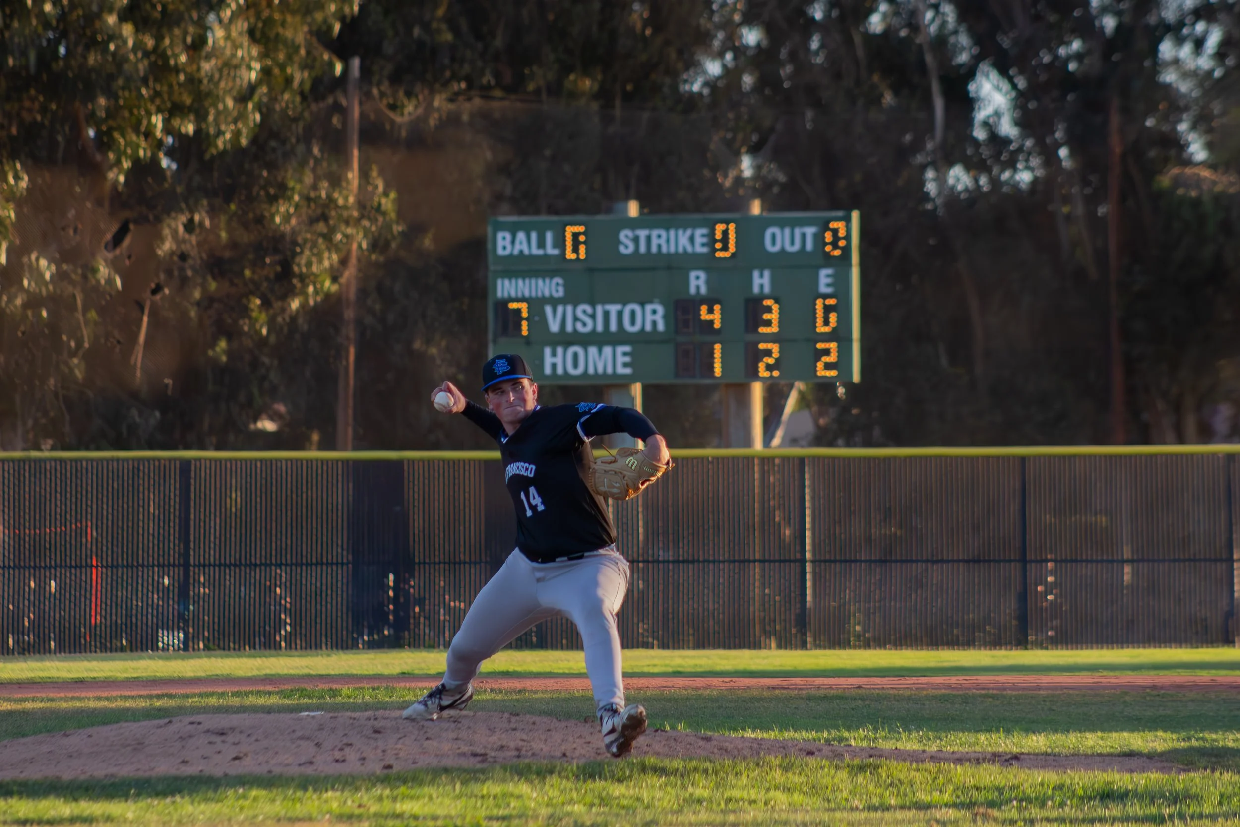 A baseball pitcher in a black jersey and gray pants throwing a pitch on a baseball field with a scoreboard and trees in the background.