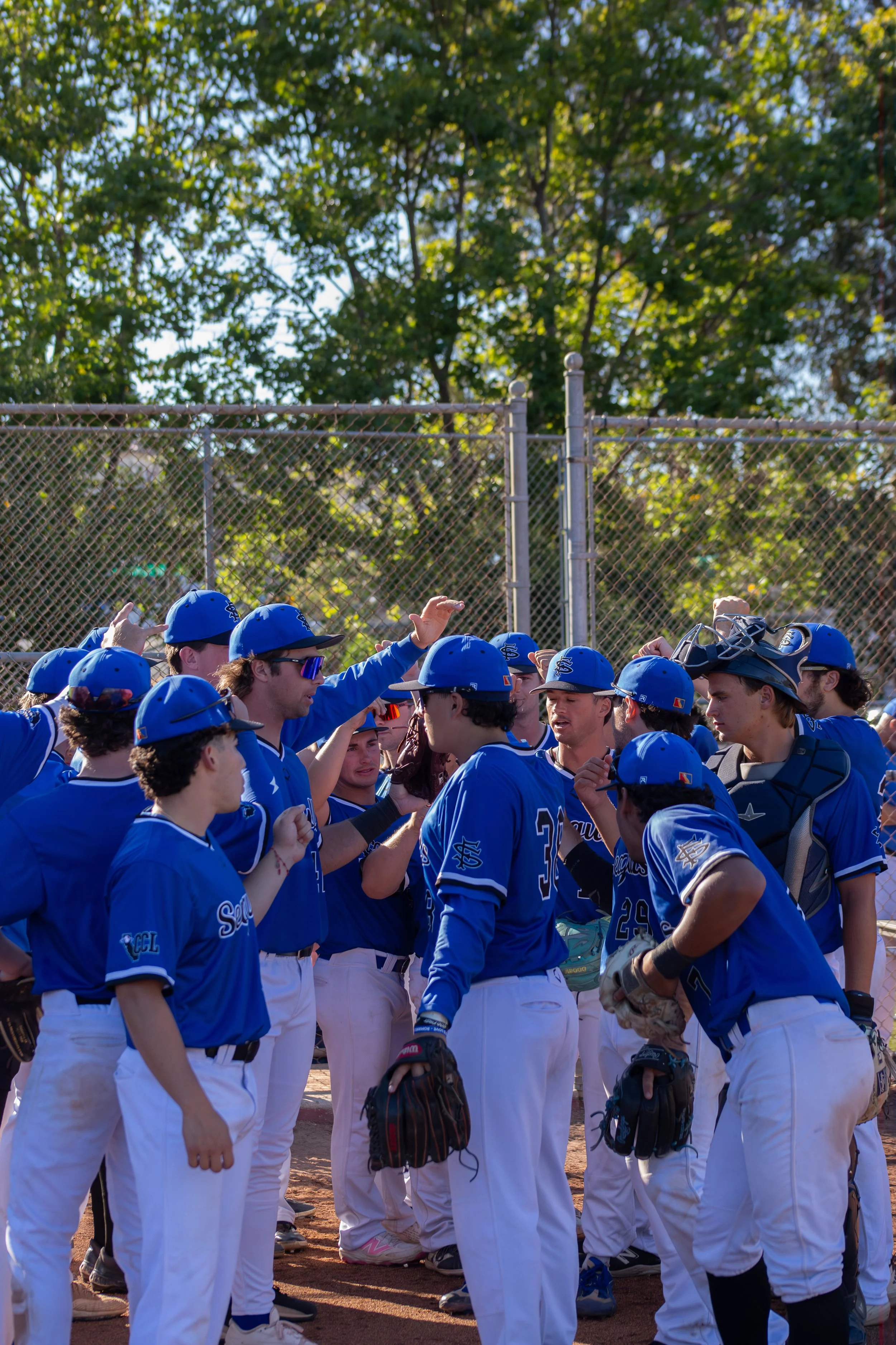 A group of young baseball players in blue jerseys and white pants gathered in a huddle on a baseball field, some wearing helmets or holding gloves, with trees and a chain-link fence in the background.