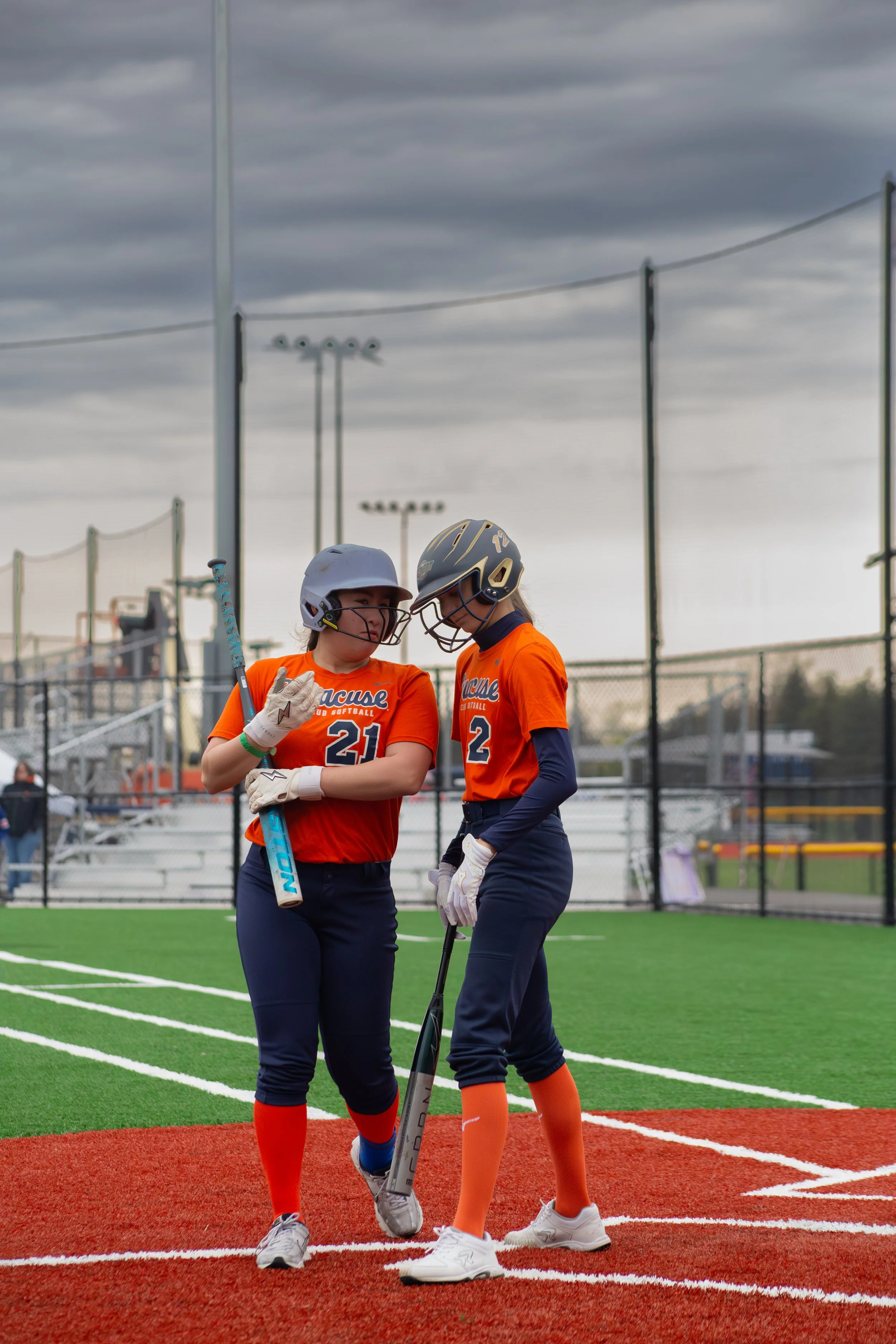 Two female softball players wearing orange and navy uniforms and helmets standing on a baseball field, talking to each other while holding a bat, with a cloudy sky overhead.