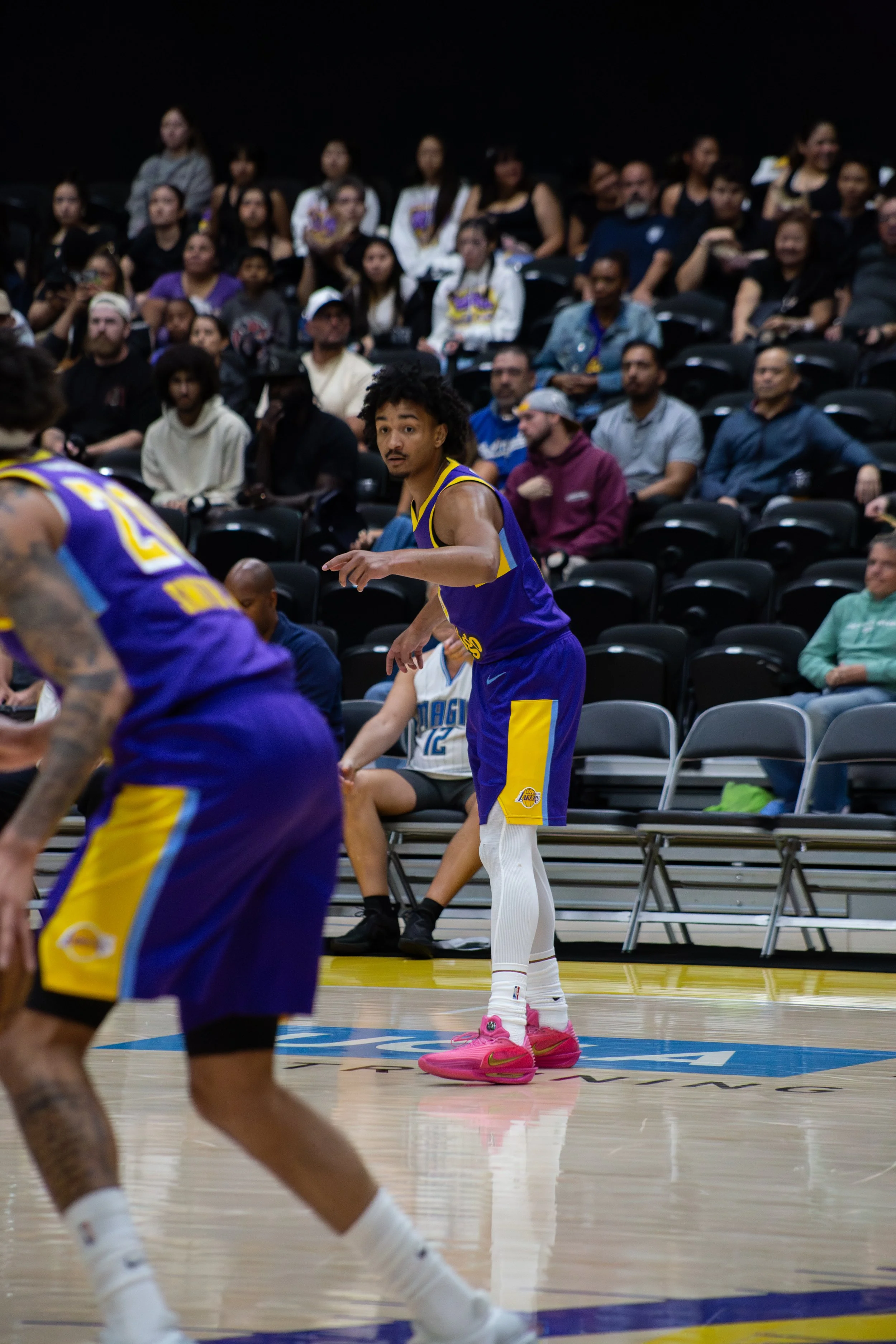 A basketball player in a blue and yellow Los Angeles Lakers uniform with pink shoes is on the court during a game with spectators sitting on black chairs in the background.
