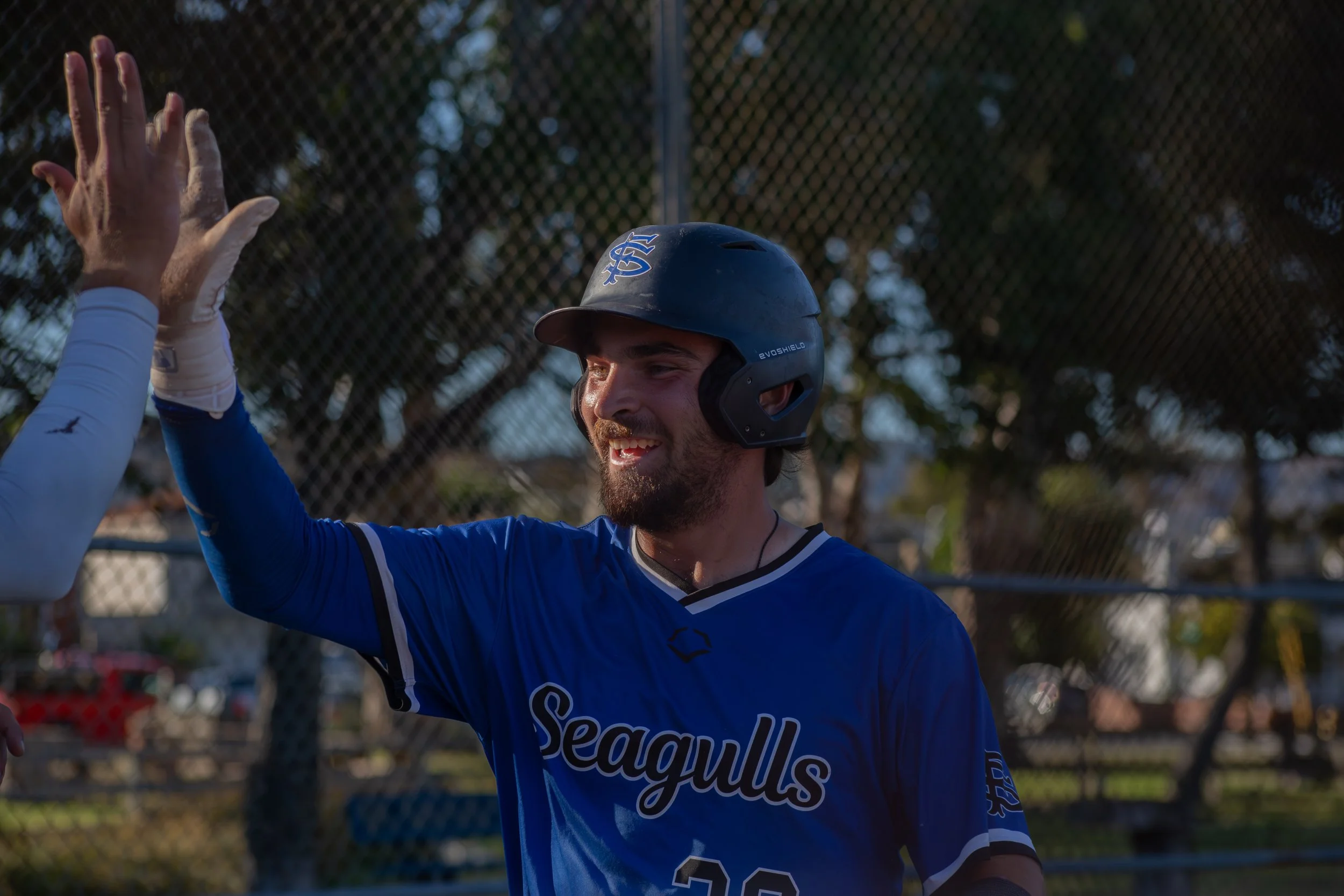 Baseball player wearing a blue jersey and helmet, smiling, giving a high five to a teammate at the baseball field.