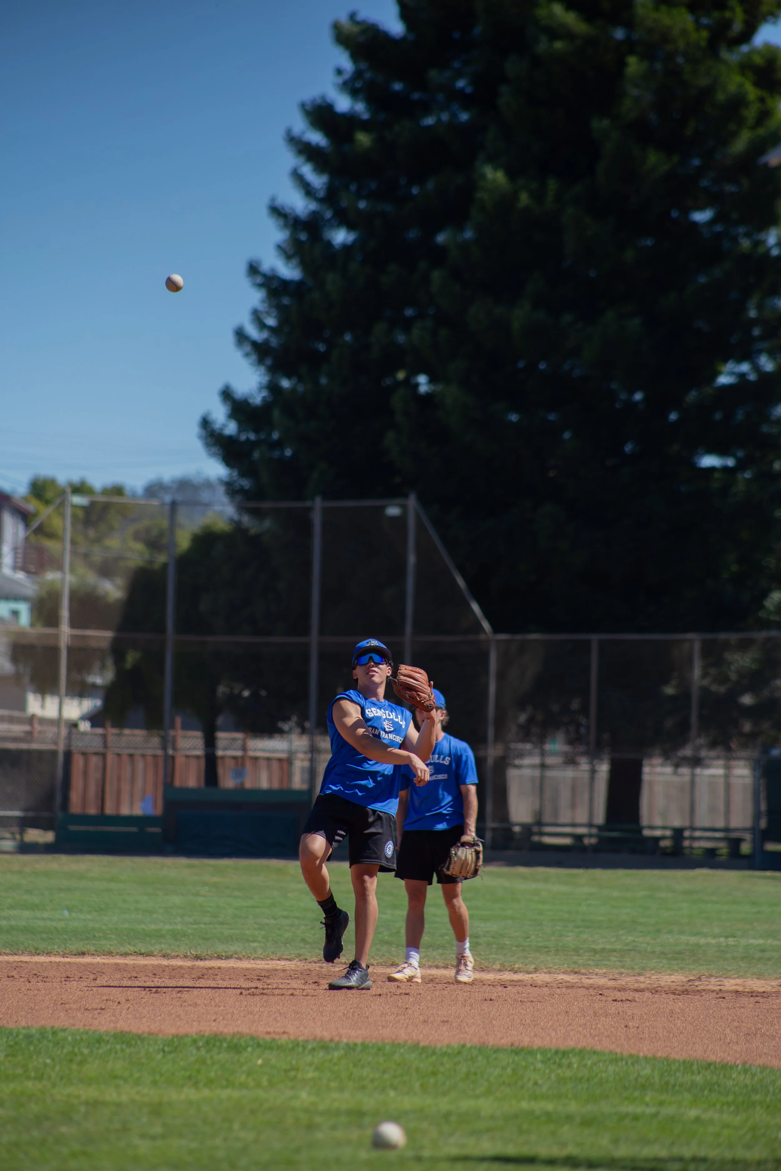 A baseball player in a blue uniform and black cleats pitches a ball during a game on a sunny day. Other players and a baseball field with a fence and trees are visible in the background.