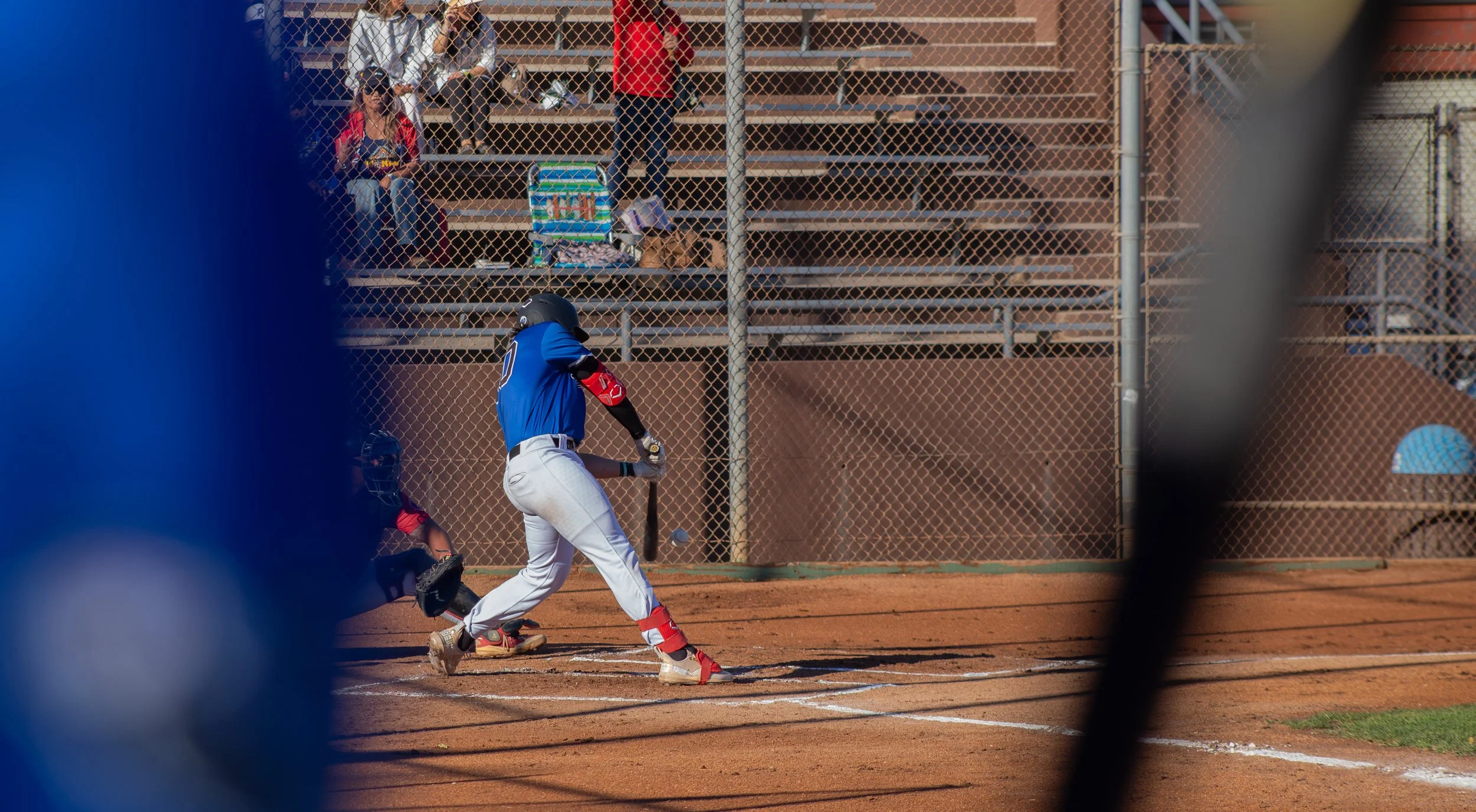 A baseball player in a blue uniform is about to hit a pitch at a baseball field, with spectators sitting in the bleachers behind a chain-link fence.