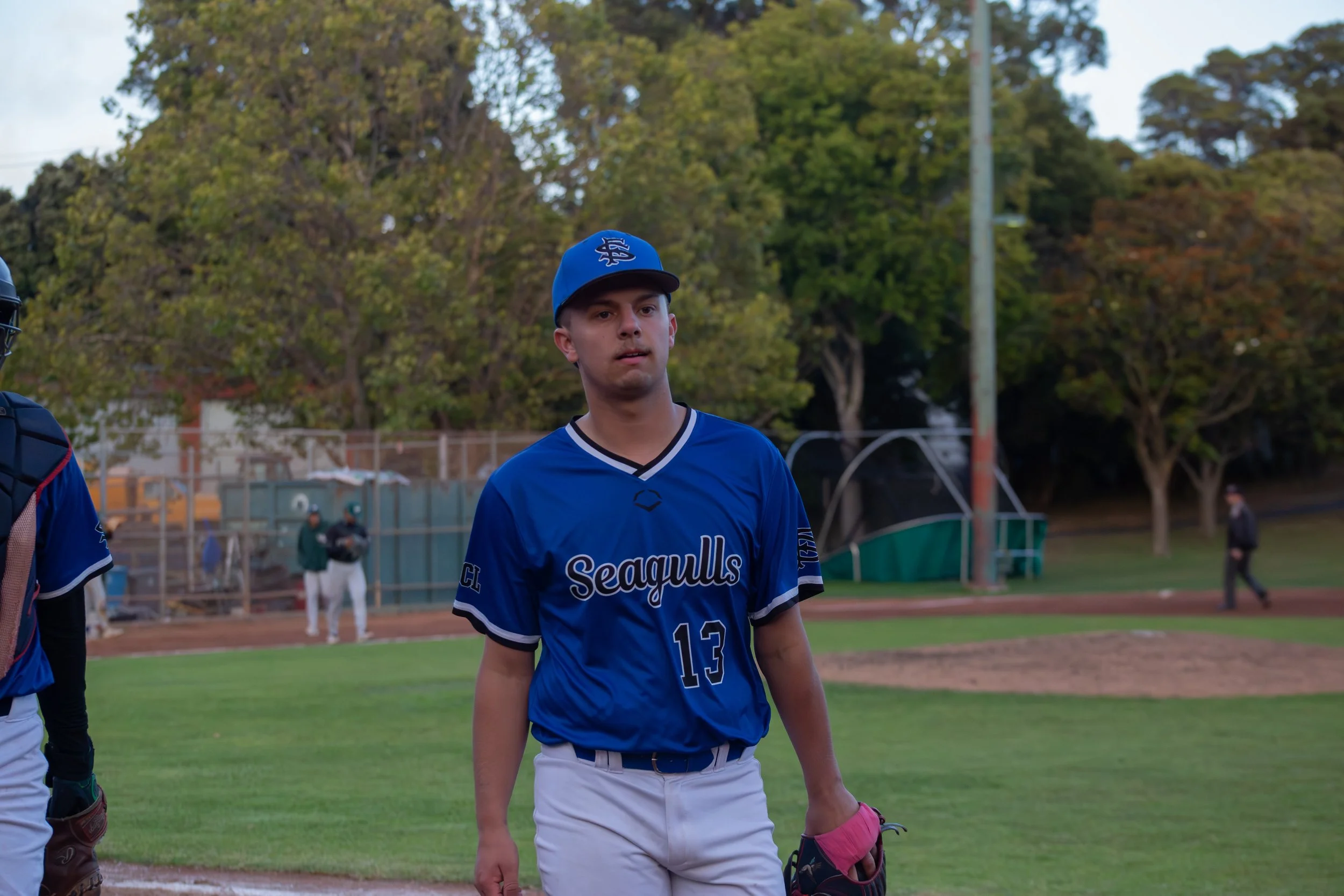 A young man in a blue baseball uniform and a blue cap stands on a baseball field. There are trees and a fence in the background, and he appears to be participating in a game or practice.