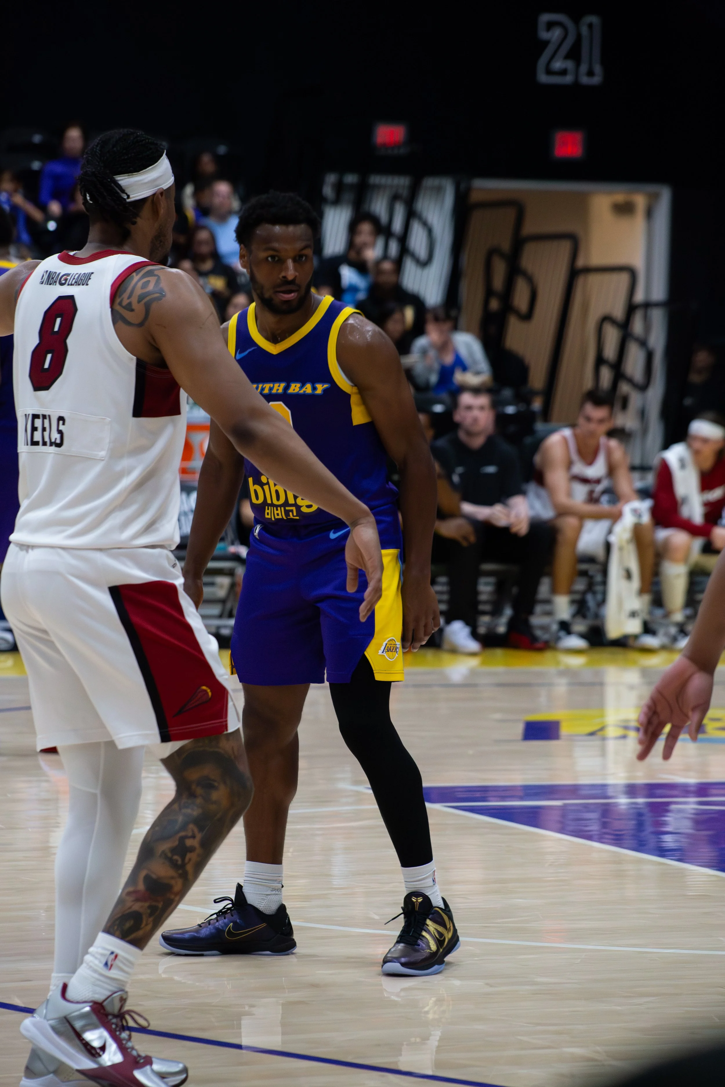 Two basketball players on the court during a game, one wearing a white jersey and the other wearing a blue and yellow jersey. The player in white has a tattoo on his arm and is wearing a headband, while the player in blue appears to be talking or dis
