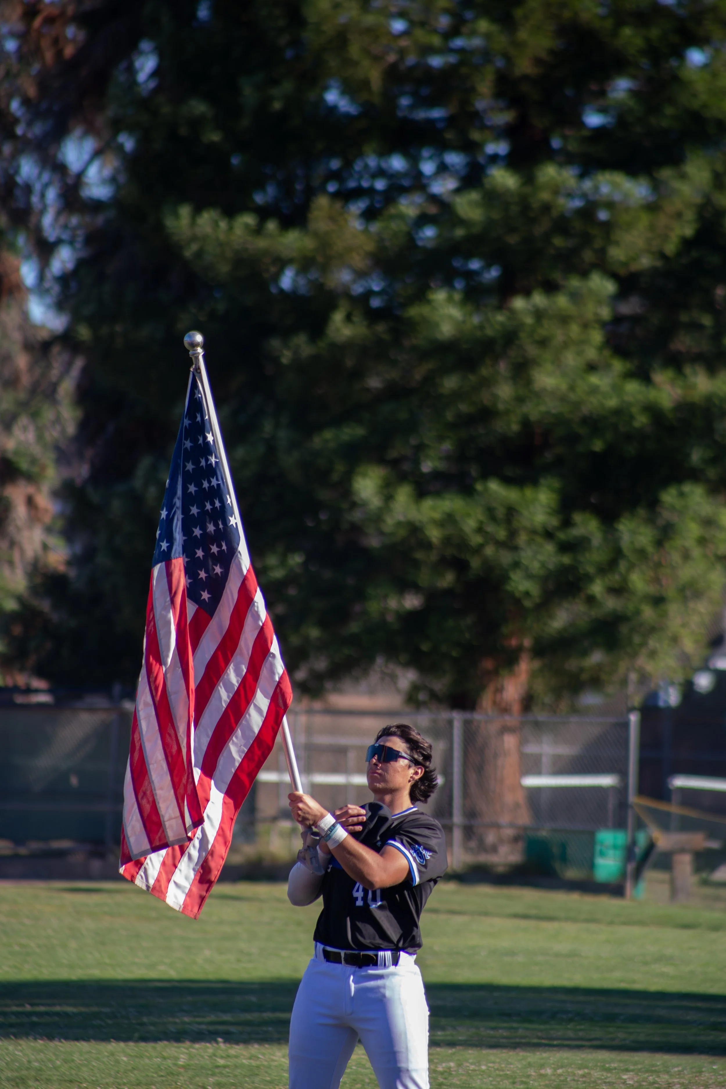 A person wearing sunglasses and a black sports jersey holding an American flag on a flagpole at a sports field with trees in the background.