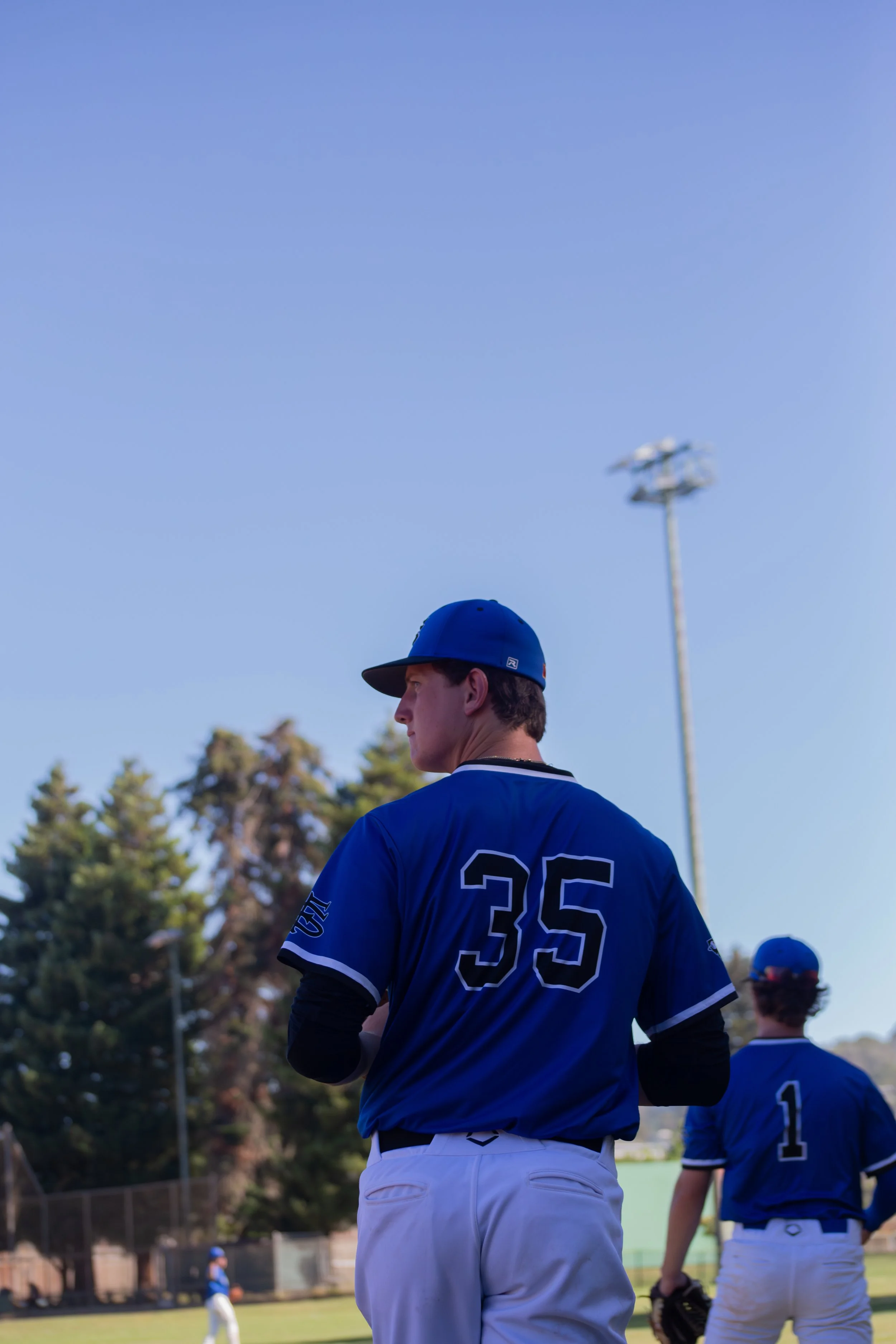 Baseball players in blue uniforms standing on the field during a game, with trees and a clear blue sky in the background.