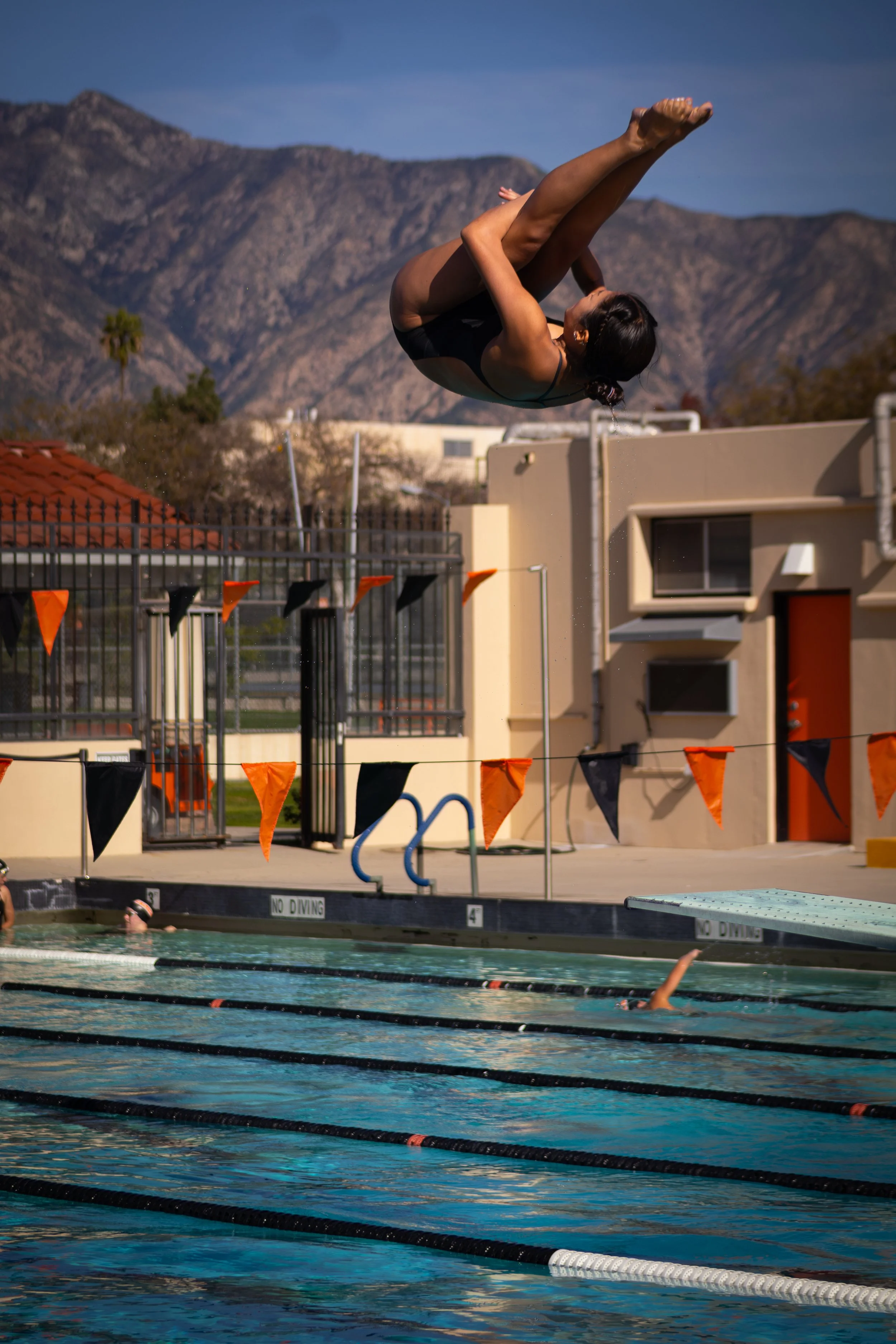 A swimmer doing a flip dive into an outdoor swimming pool with lane markers, surrounding fencing, and a building, with mountains in the background under a clear sky.