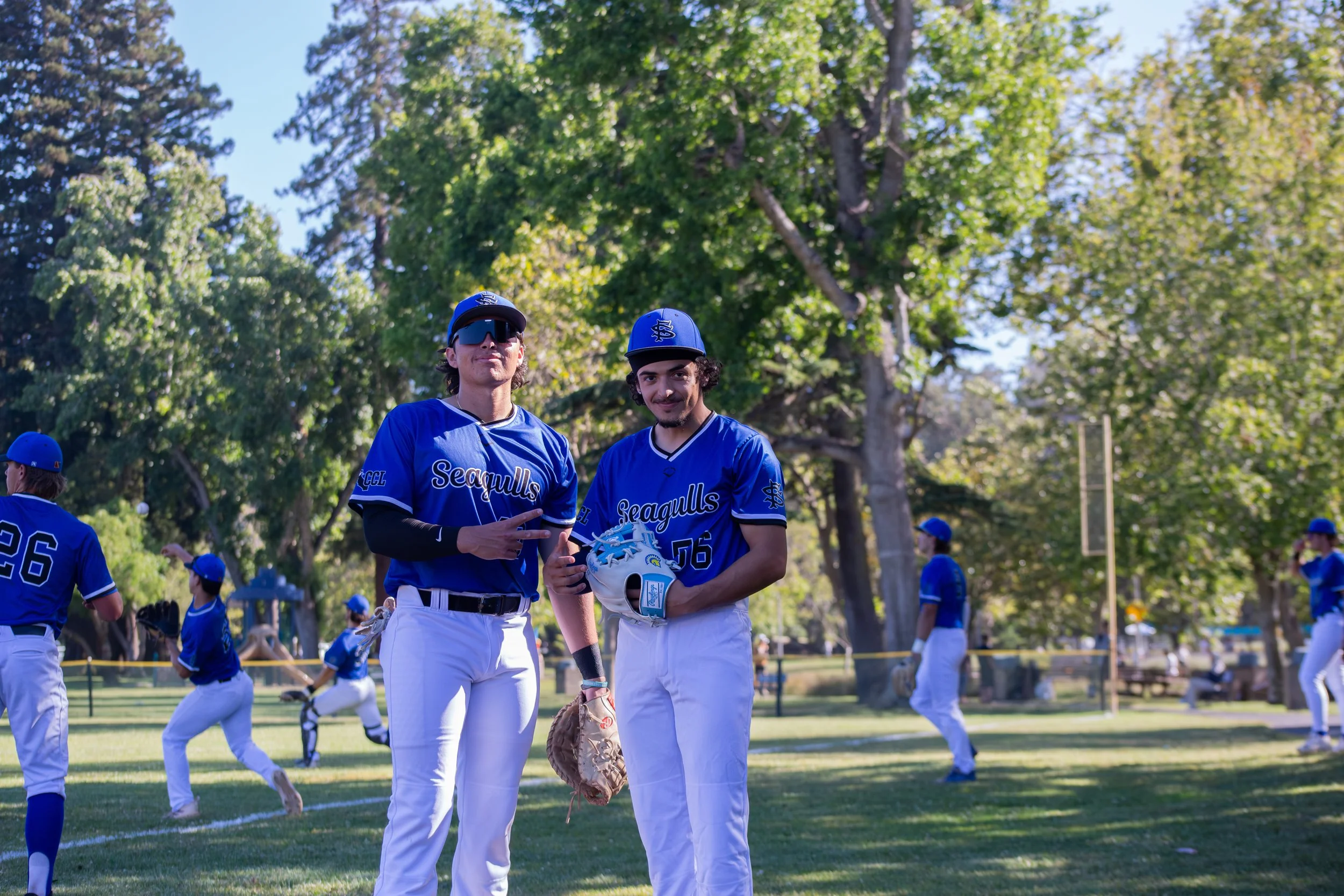 Two baseball players in blue uniforms standing on a grassy field with other players and trees in the background.