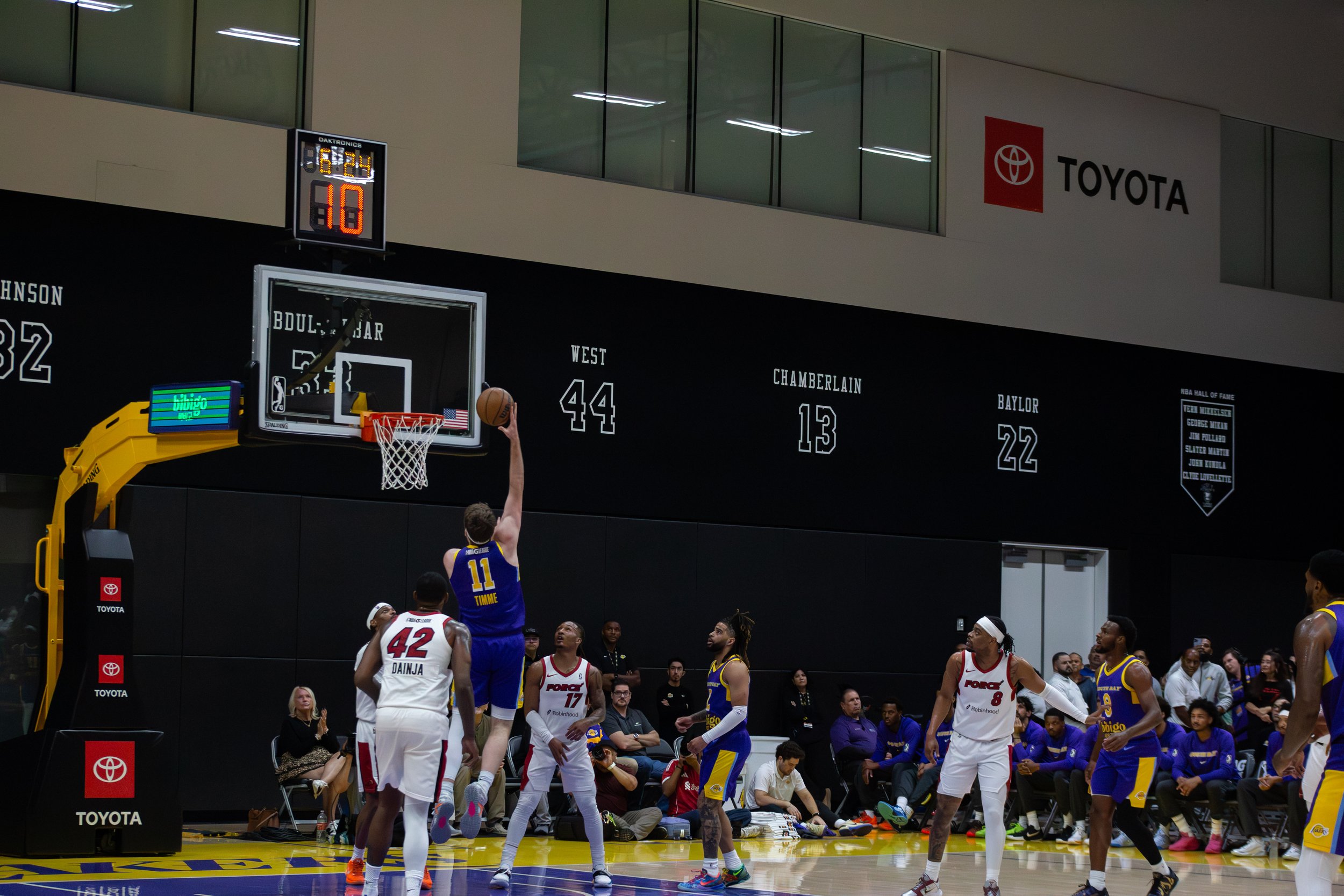 A basketball game in progress with players near the basket. One player in blue jersey number 11 is taking a shot. Opposing team players in white jerseys are defending. Spectators and team staff observe from the sidelines. Players are on a basketball 