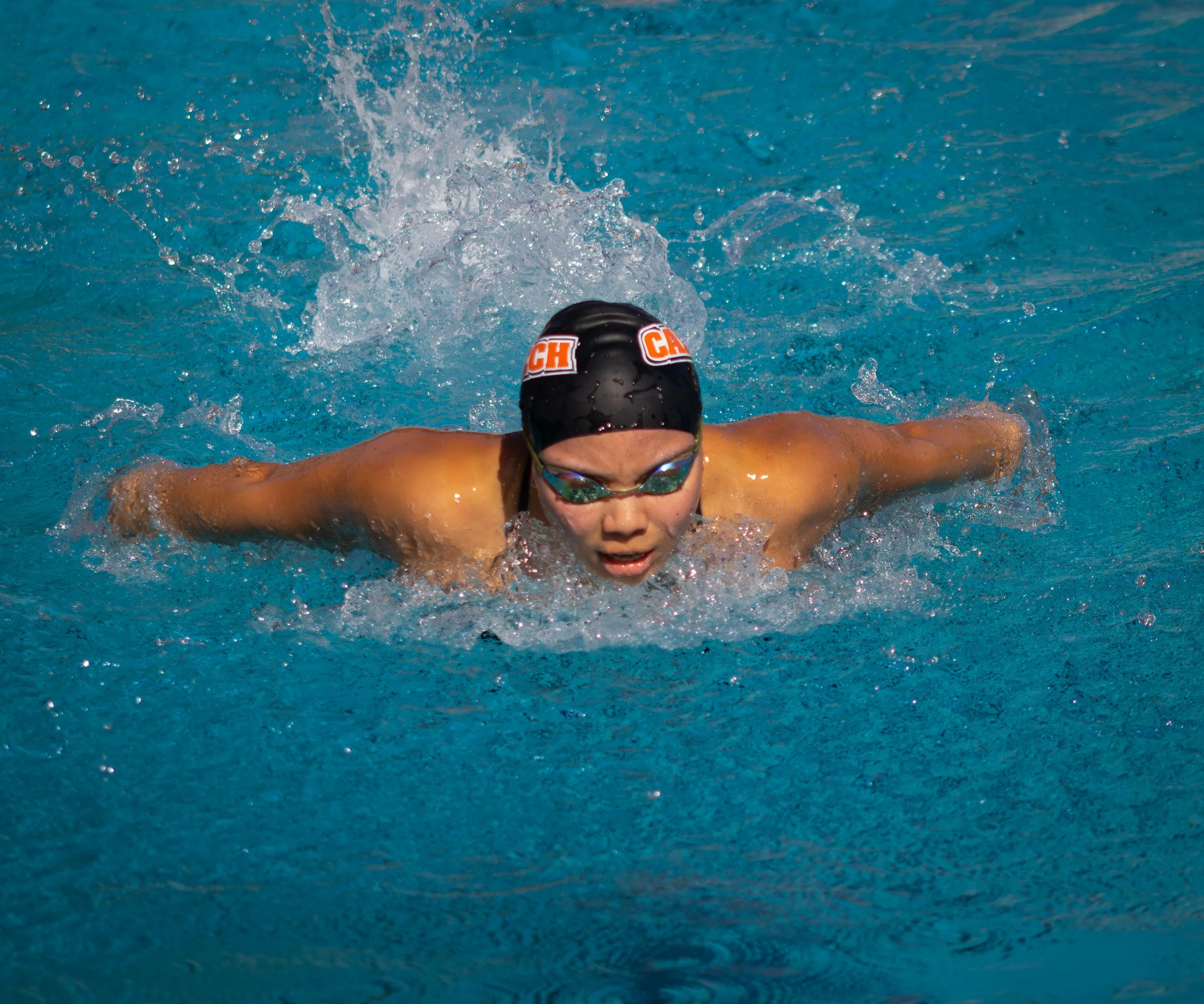 Swimmer performing the butterfly stroke in an outdoor swimming pool, wearing a black swim cap with red and white branding, goggles, and swimming in clear blue water.