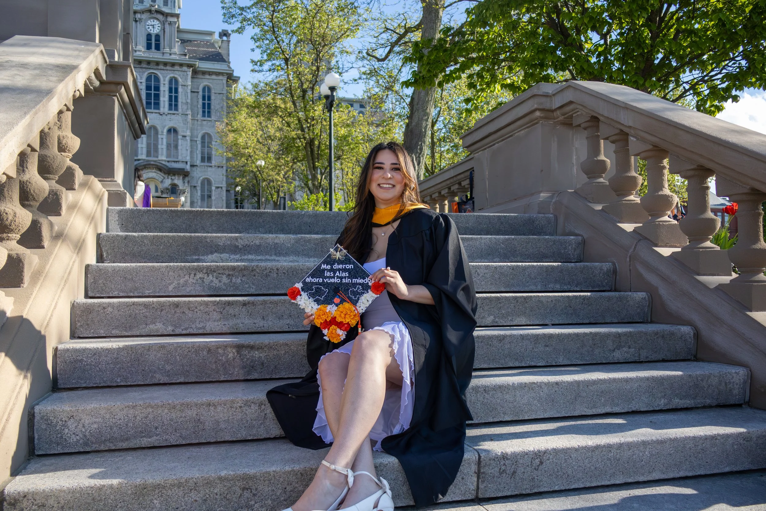 A young woman in a graduation cap and gown sitting on stone steps outdoors, holding a decorated graduation cap, with a historic building, trees, and a clear sky in the background.