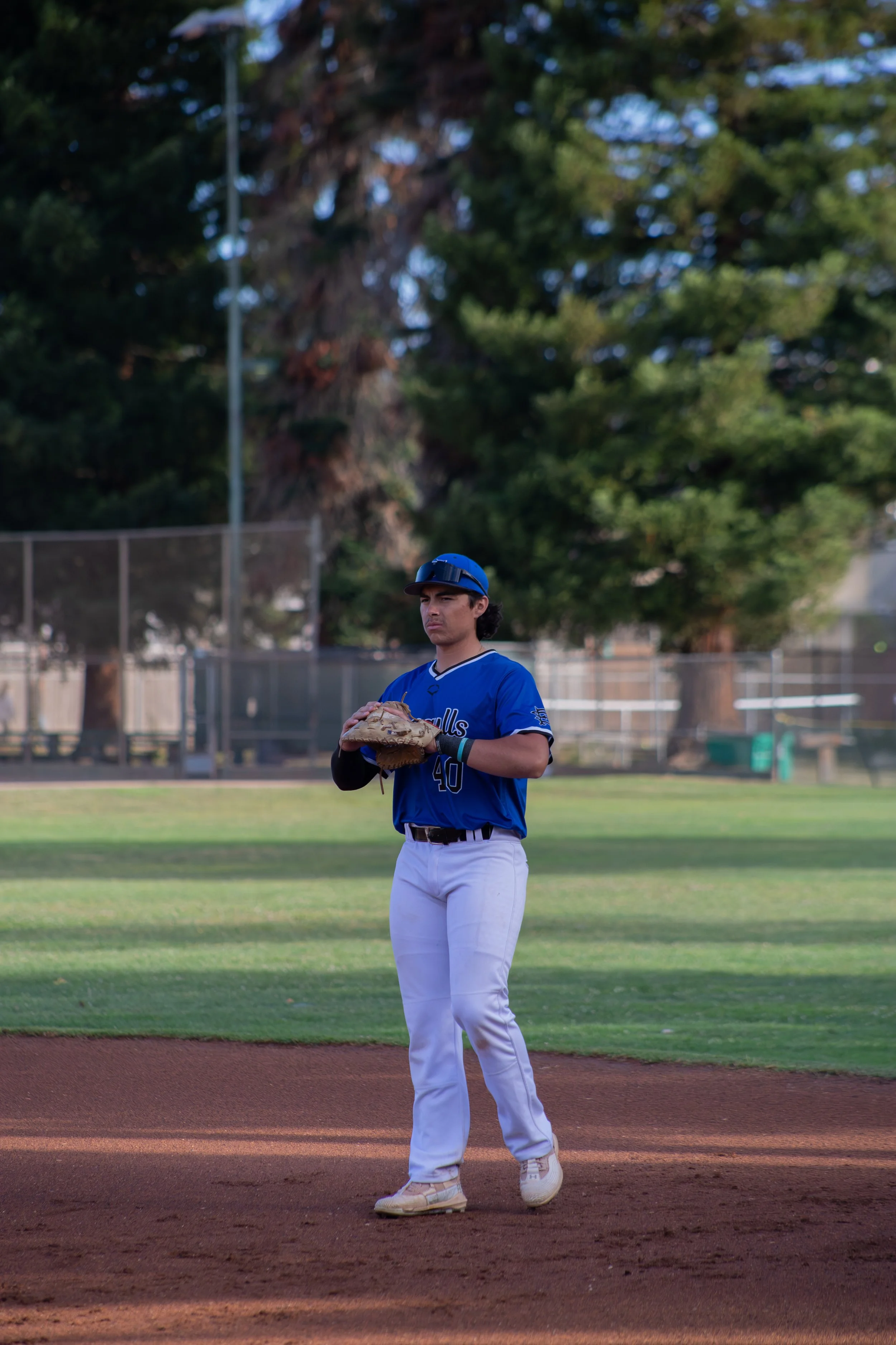 A young baseball player wearing a blue jersey, white pants, and a protective helmet stands on the infield of a baseball field holding a baseball glove. In the background, there are tall trees and a chain-link fence surrounding the field.
