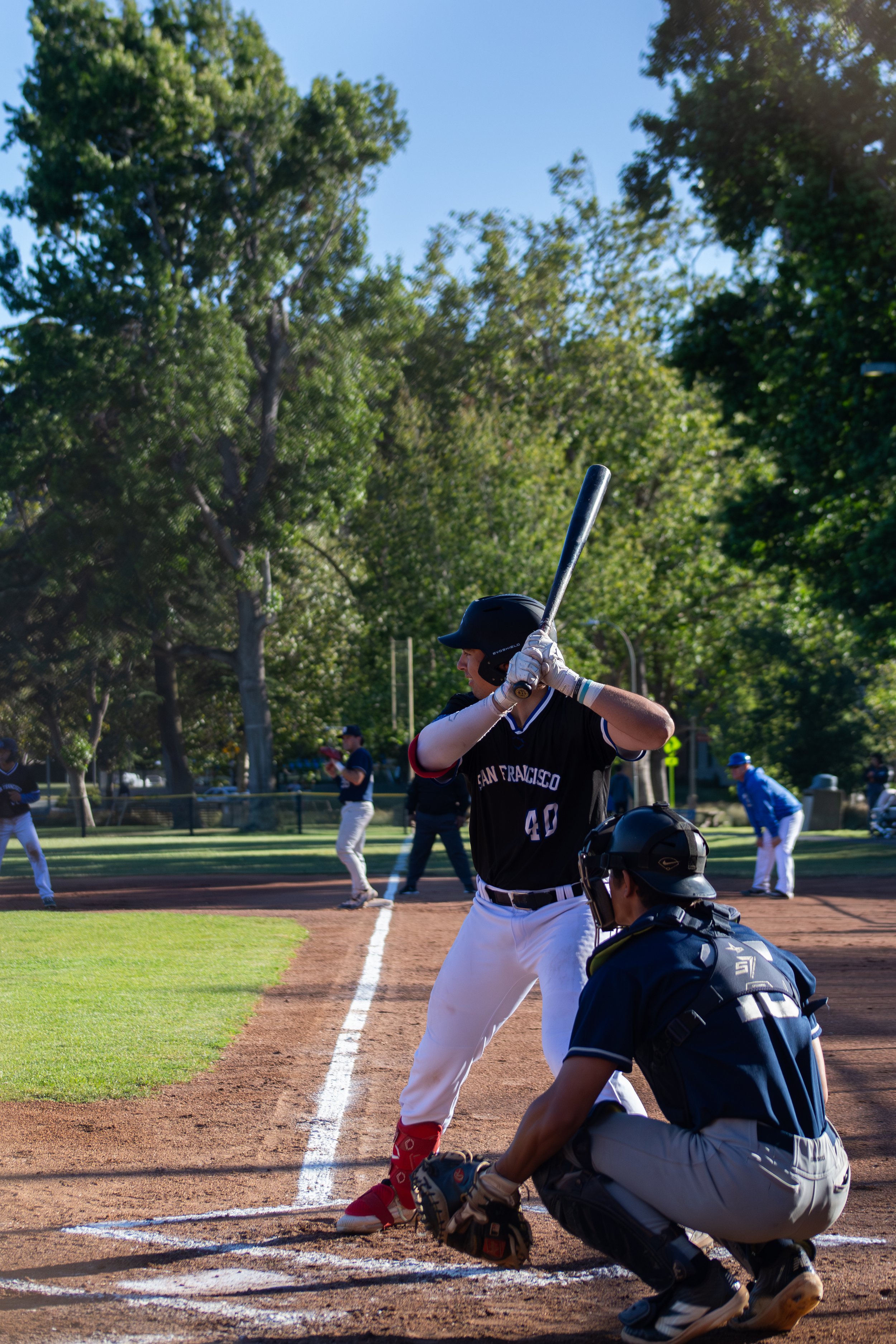 A baseball game with players at bat, pitcher, and catcher on a sunny day in a park with trees in the background.