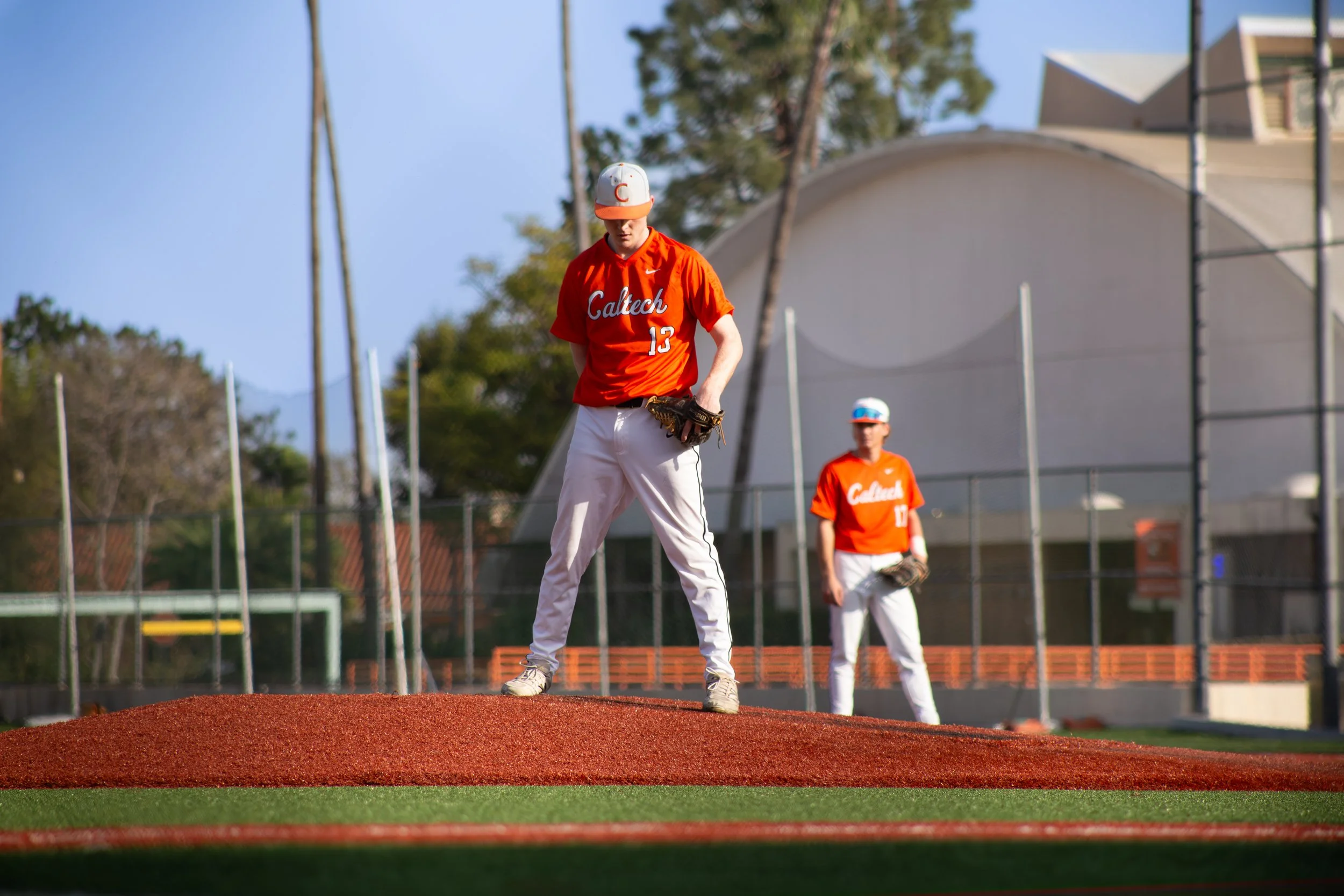 A baseball player wearing a red Caltech jersey with the number 13 and white pants is standing on a mound of dirt on a baseball field, looking down while holding a glove. Another player in a similar uniform is seen in the background, looking away. The