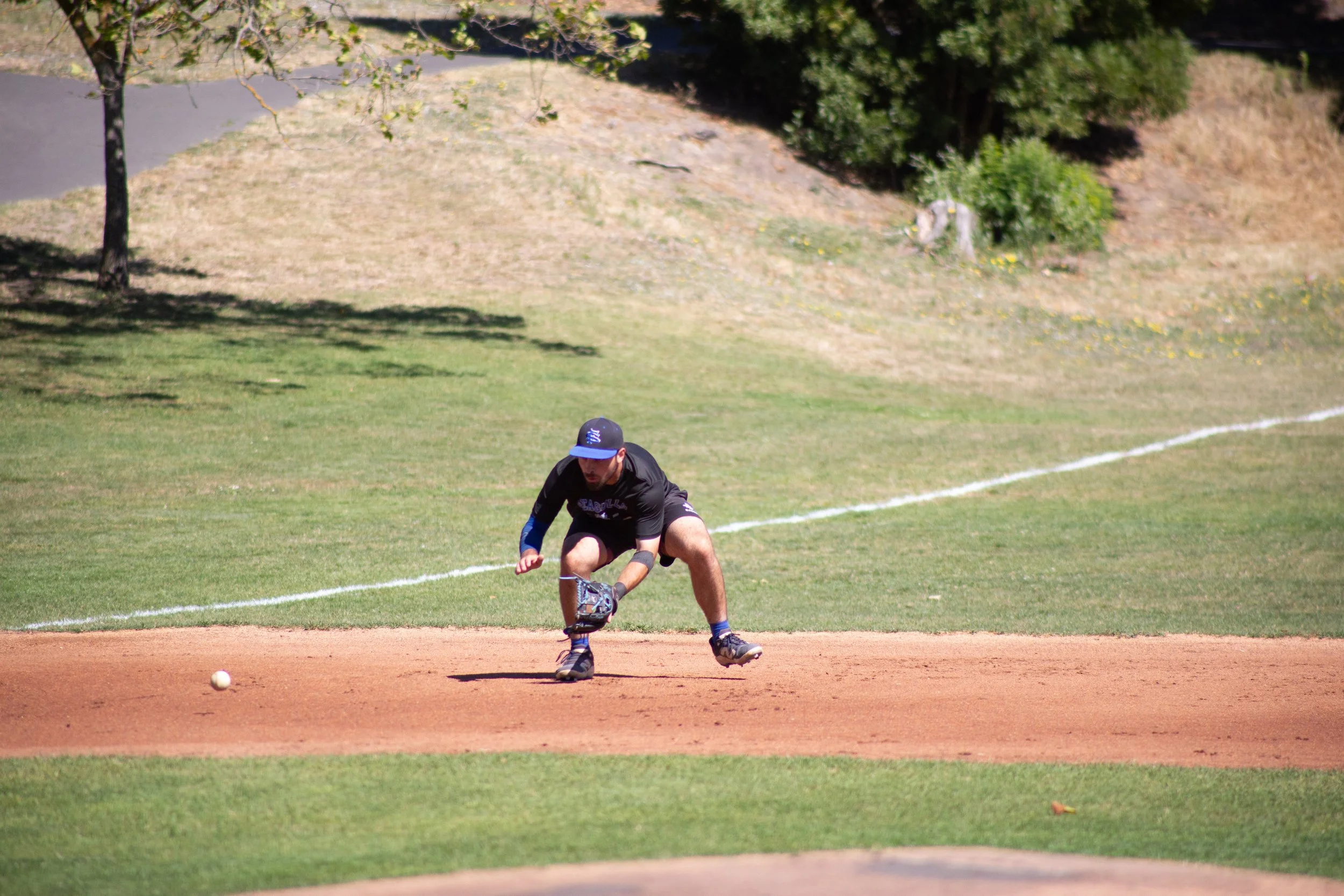 A man playing baseball on a field, fielding a ball near the pitcher's mound with grass, dirt, trees, and a small hill in the background.