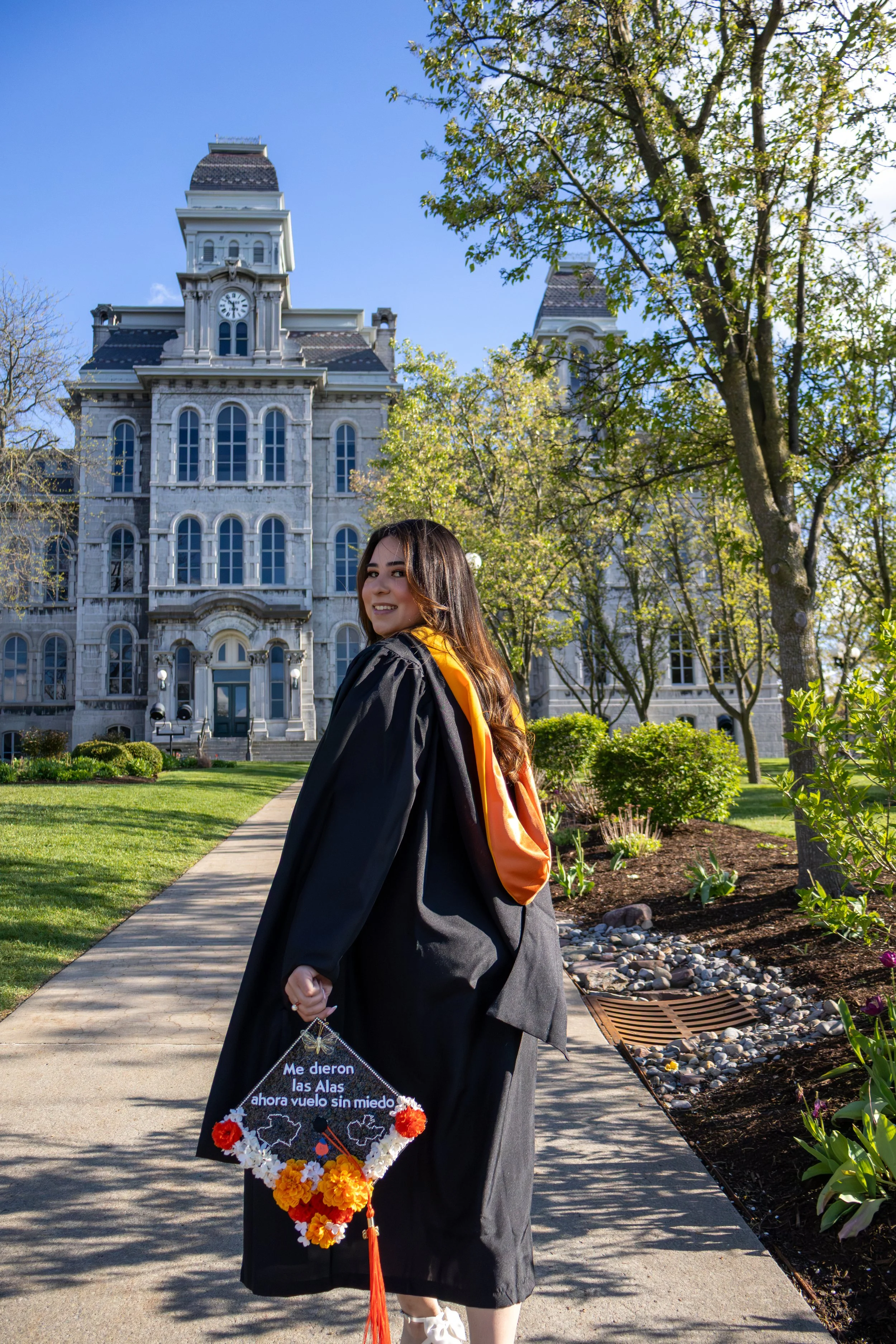 A young woman in a graduation gown holds a decorated diploma cover with flowers, standing on a sidewalk with a historic building and trees in the background on a sunny day.