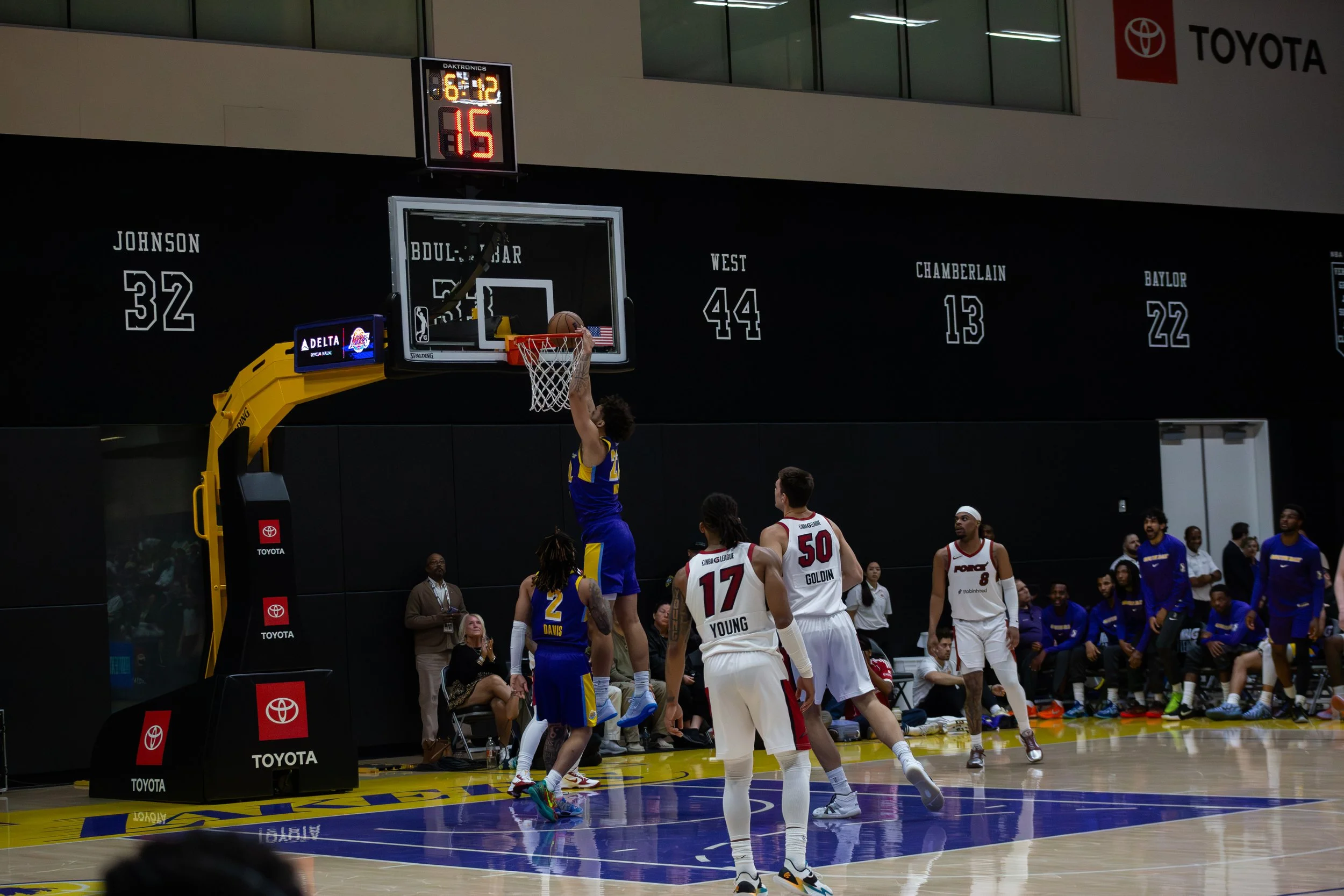 A basketball game in progress with players on the court, some shooting and others defending. The scoreboard shows 6:12 remaining on the game clock and a score of 15. Players wear uniforms with numbers and team names. Spectators and officials are watc