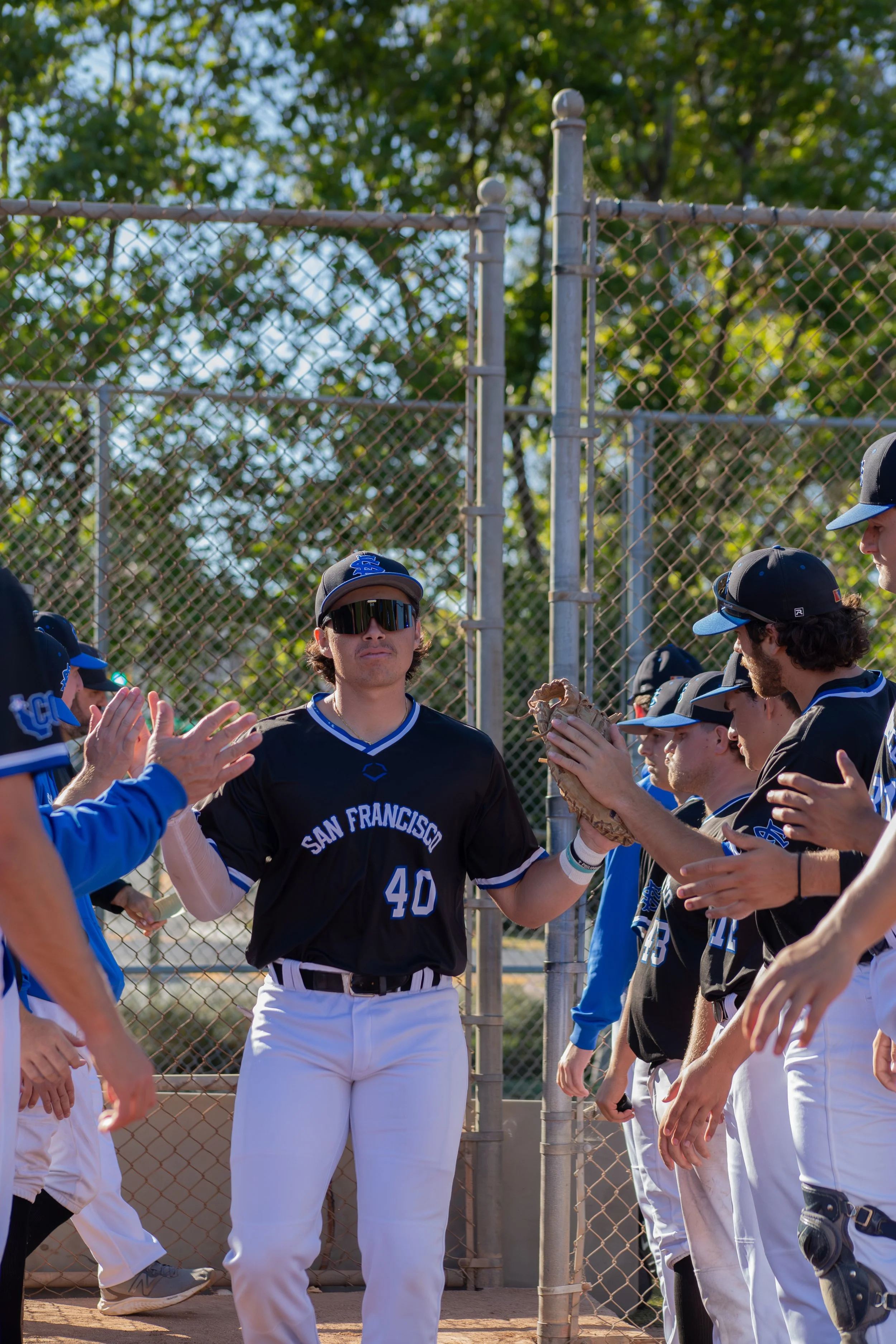 Baseball players in uniforms and caps, some with sunglasses, forming a line for a processional at a baseball field, with a chain-link fence and green trees in the background.