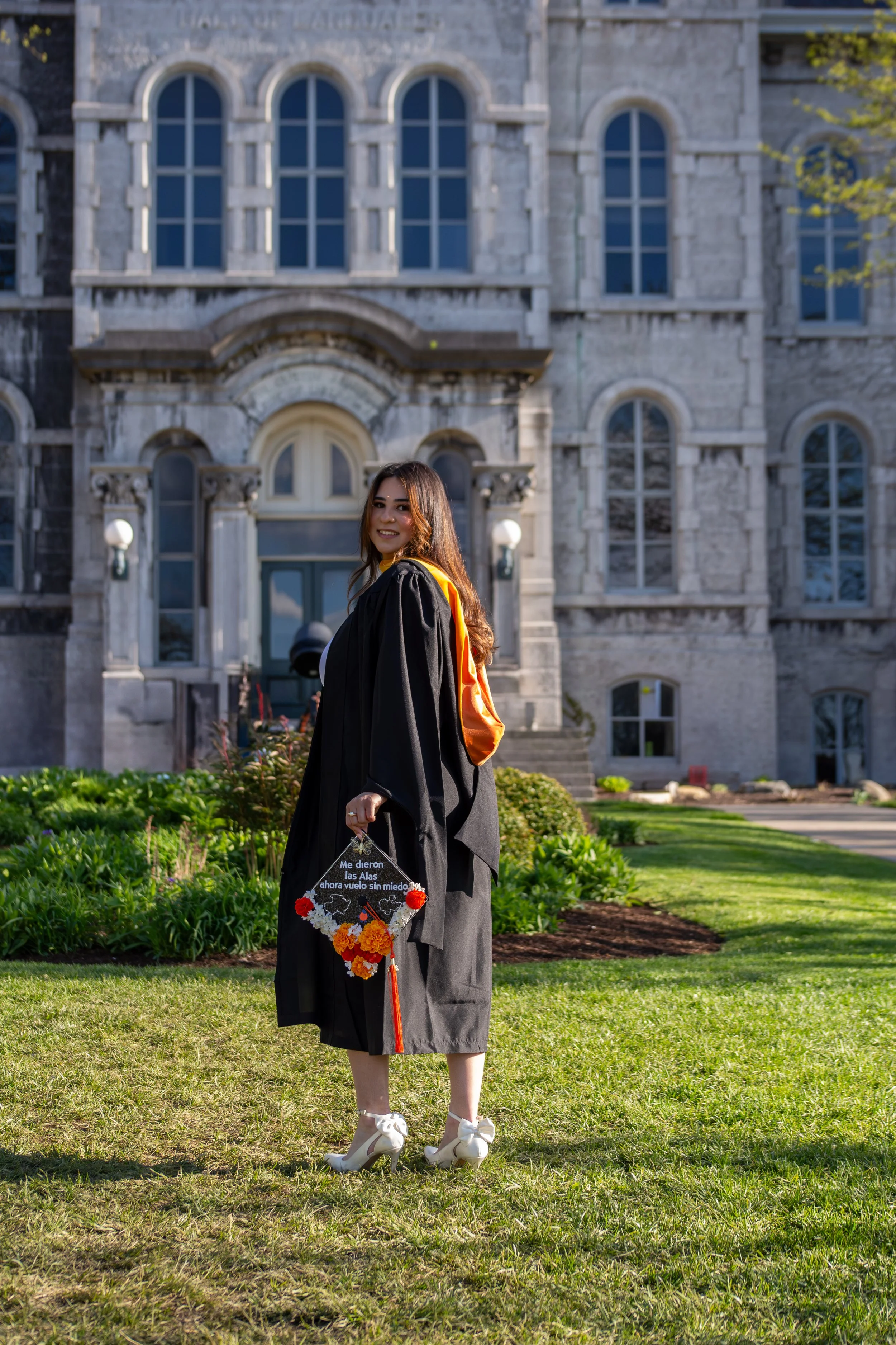 Young woman in a graduation cap and gown holding a decorated graduation cap with flowers stands on a grassy area in front of a large stone building with arched windows.