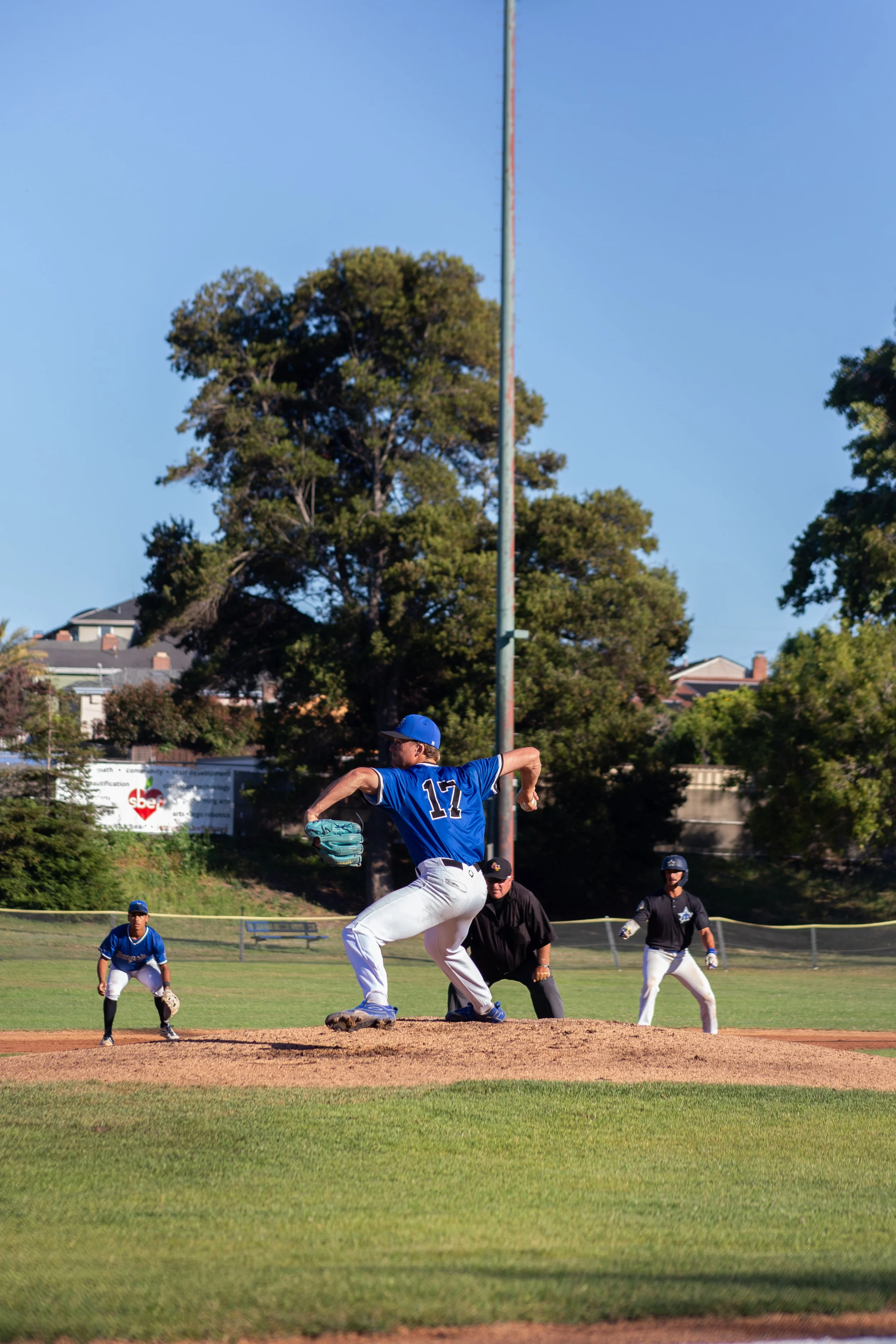 A baseball player wearing a blue jersey with the number 17, white pants, and a blue cap, is pitching on a baseball mound during a game. Other players are visible in the background on the field, along with two spectators and trees in the distance.