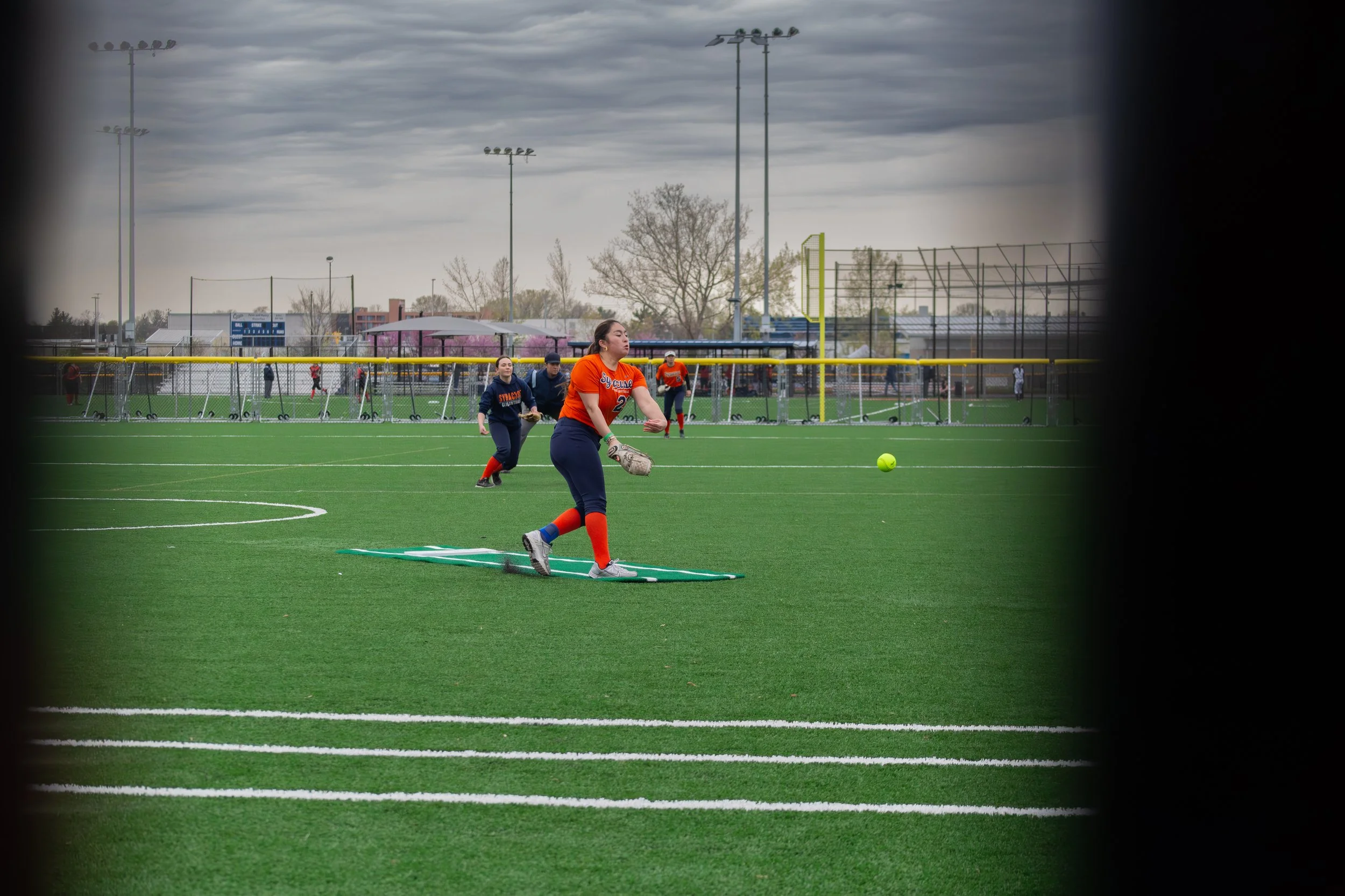 A softball team warming up on a field on a cloudy day, with players stretching and practicing near the dugout, seen through a fence.