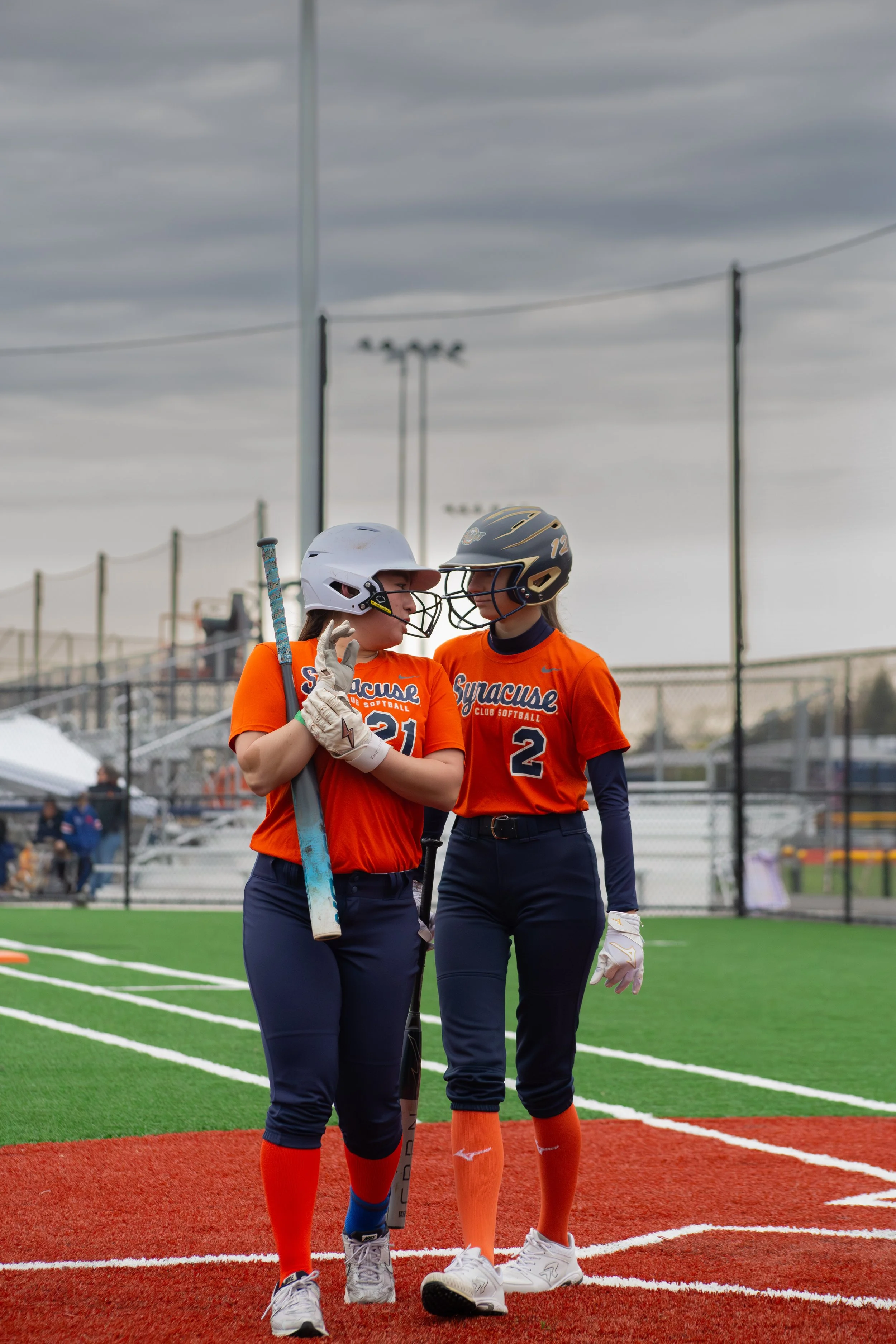 Two female softball players wearing orange jerseys and navy pants, one wearing helmet and holding a bat, talking on a softball field under a cloudy sky.