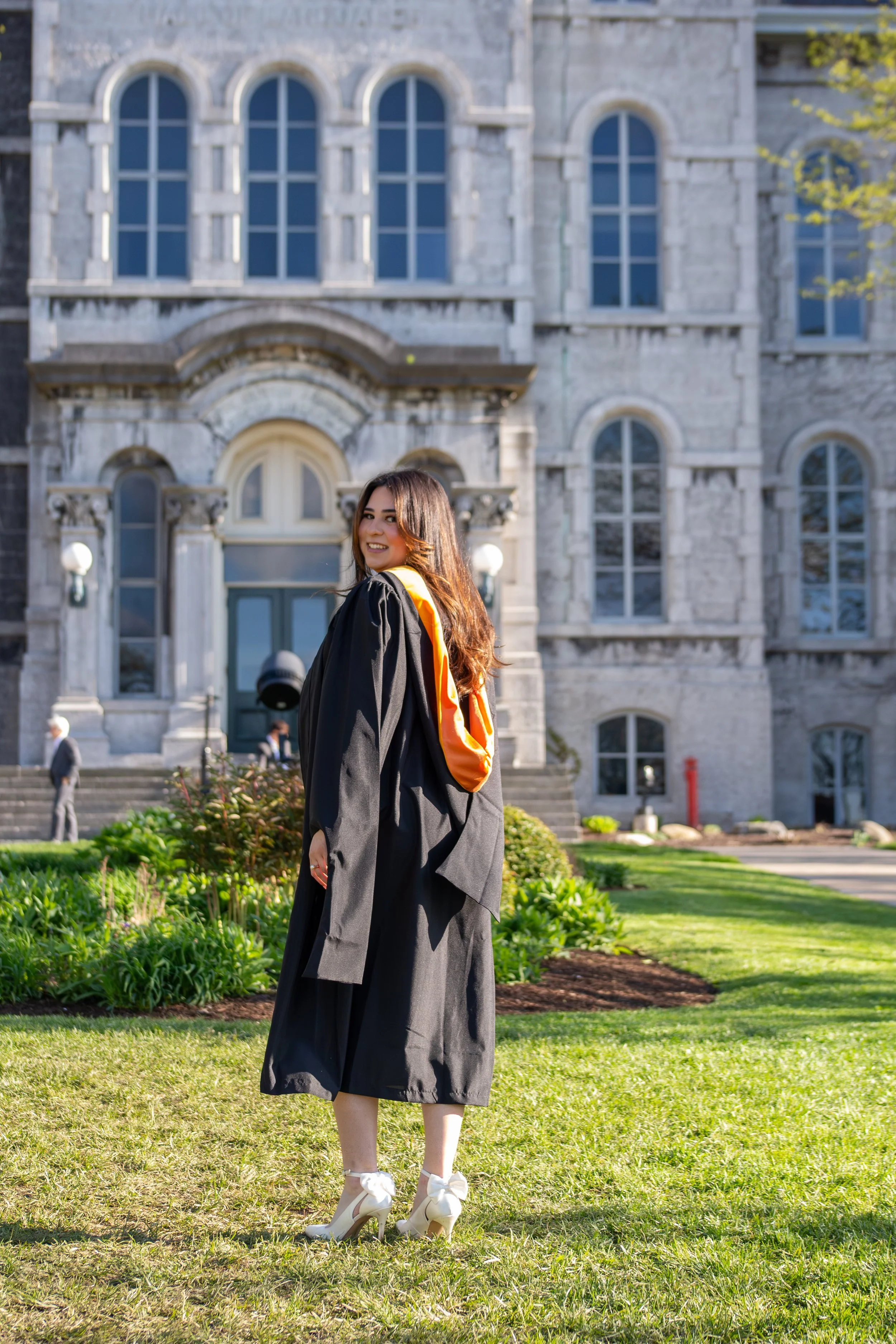 Young woman in a graduation cap and gown standing on grass in front of a historic stone building with large arched windows, smiling at the camera.