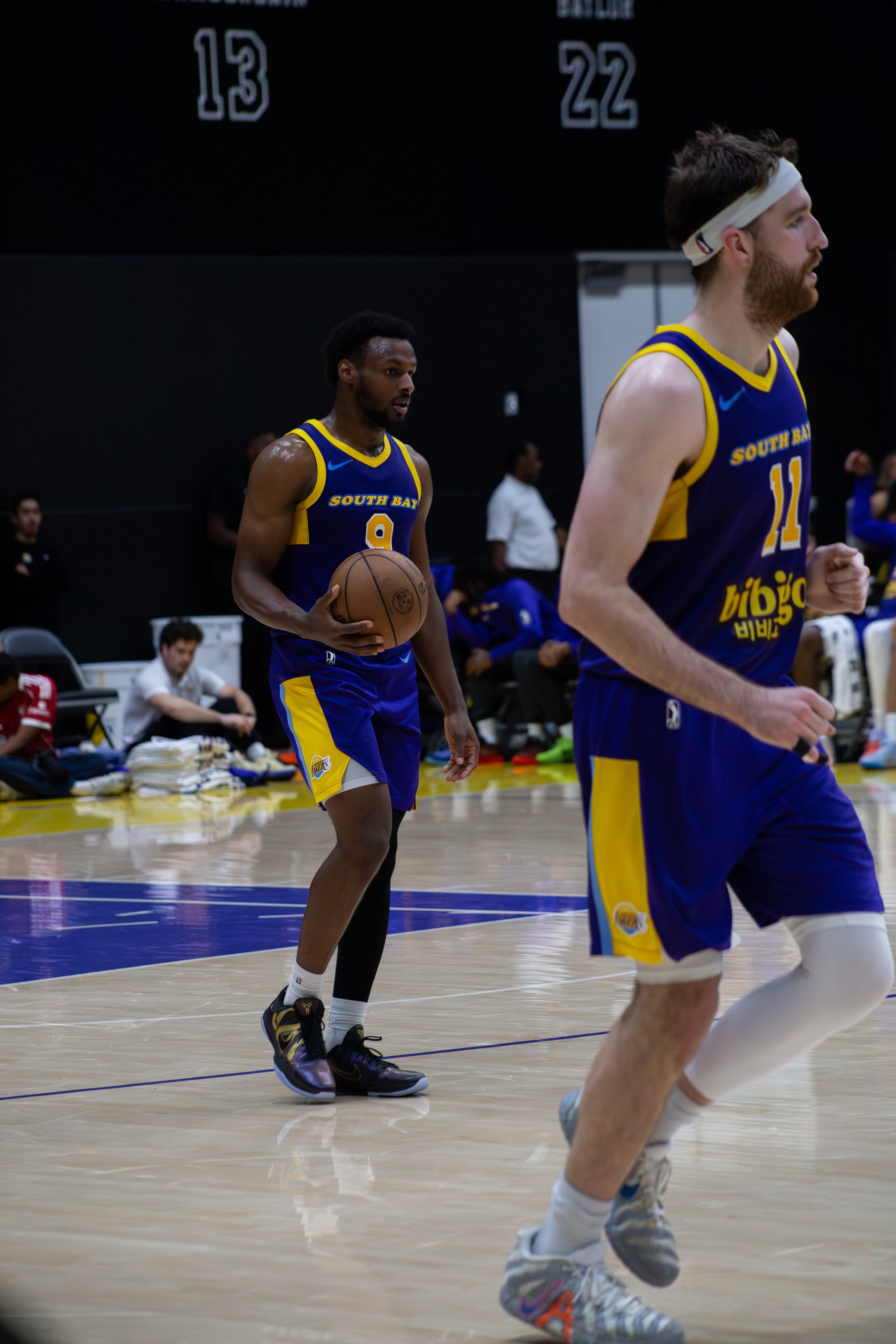 Two basketball players in yellow and blue jerseys with 'South Bay' written on them, one holding a basketball, are warming up on a basketball court. The player in the foreground is running with others also visible in the background.