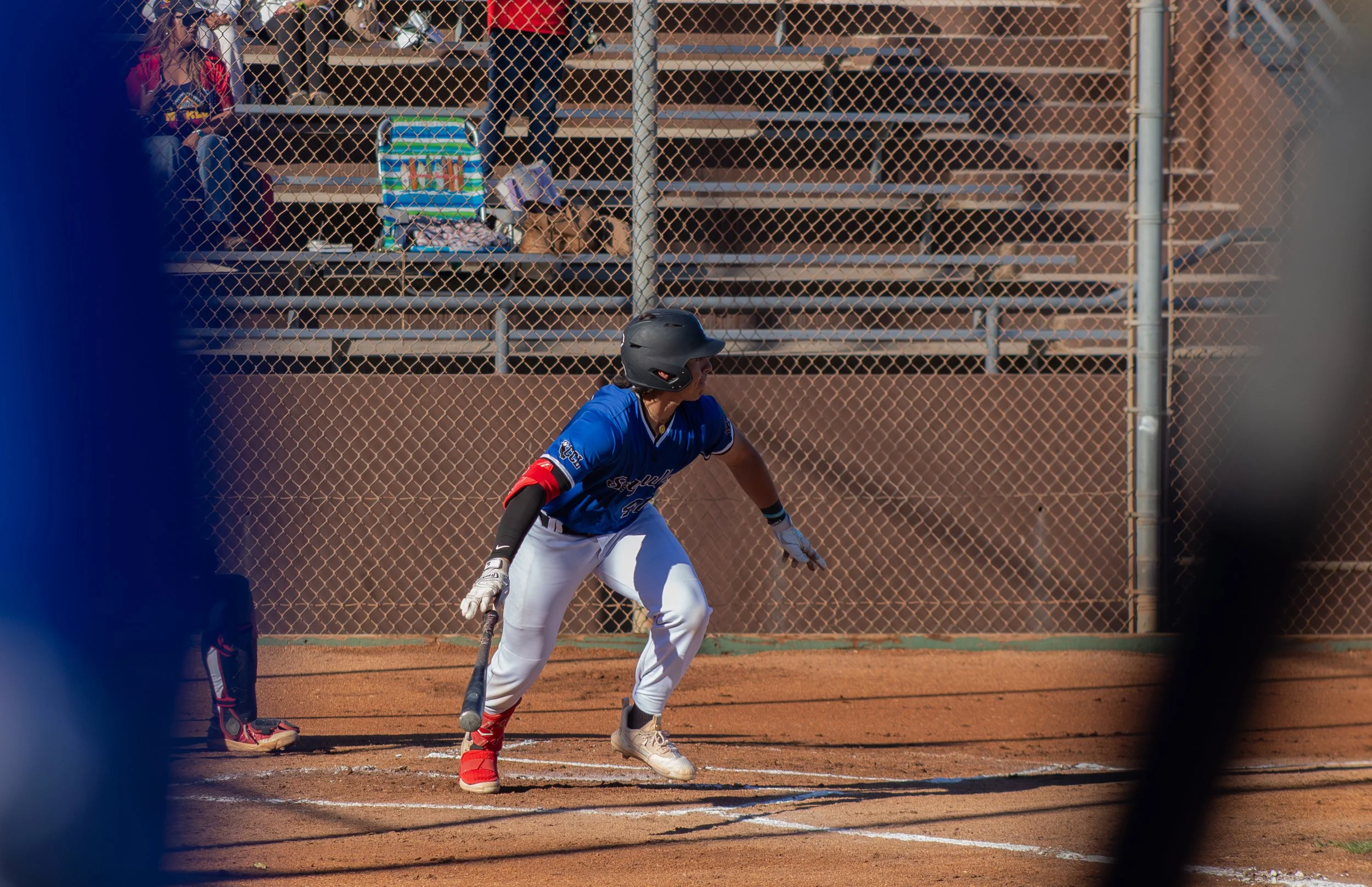A baseball player in a blue uniform and black helmet is in a batting stance on a baseball field. The field has a dirt infield with white chalk lines. Behind the player, there are empty metal bleachers and a chain-link fence. Two spectators are sittin