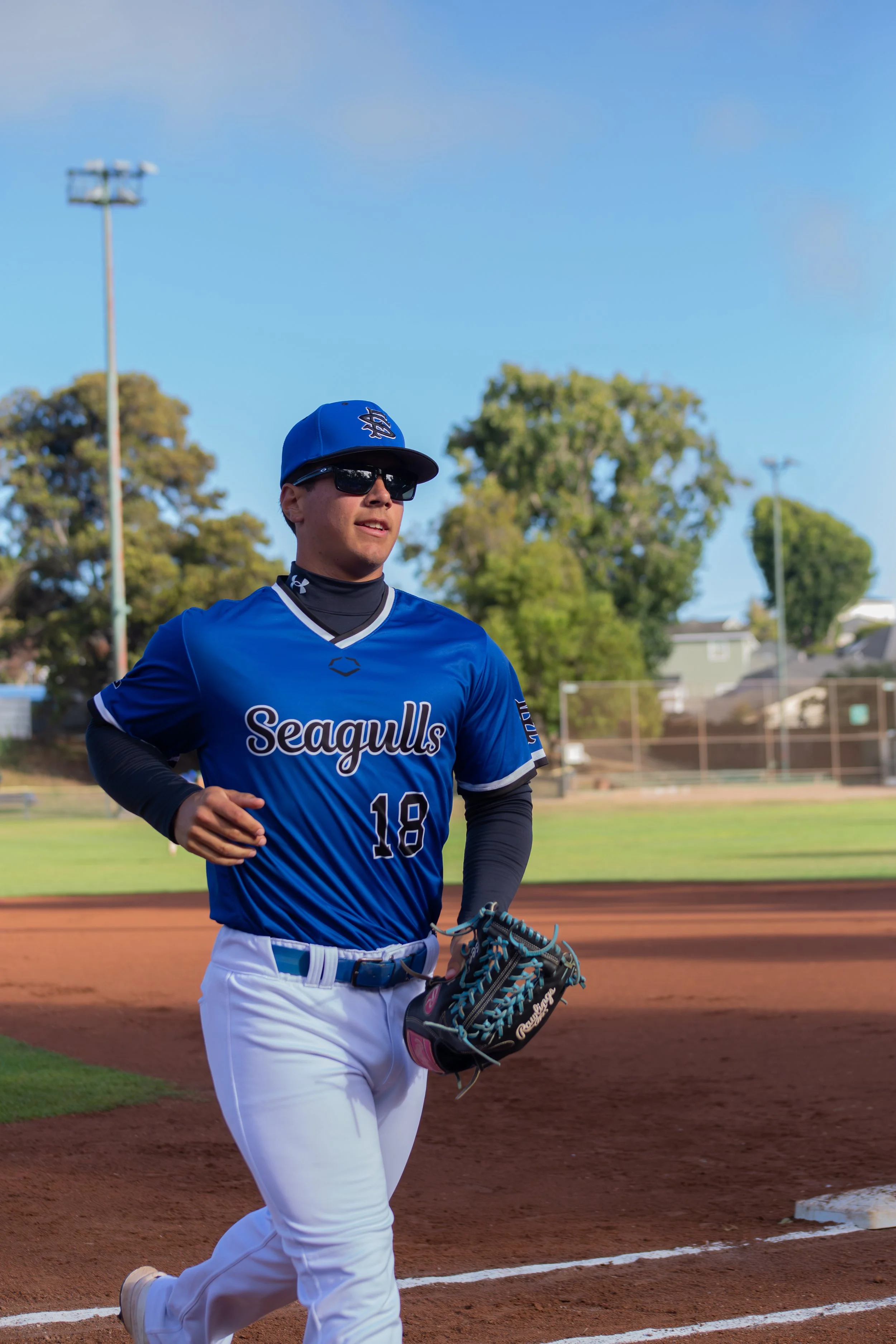 A baseball player wearing a blue jersey with 'Seagulls' and the number 18, sunglasses, and a blue cap is running on a baseball field with a glove in his left hand. There are trees, a fence, and light poles in the background under a blue sky.