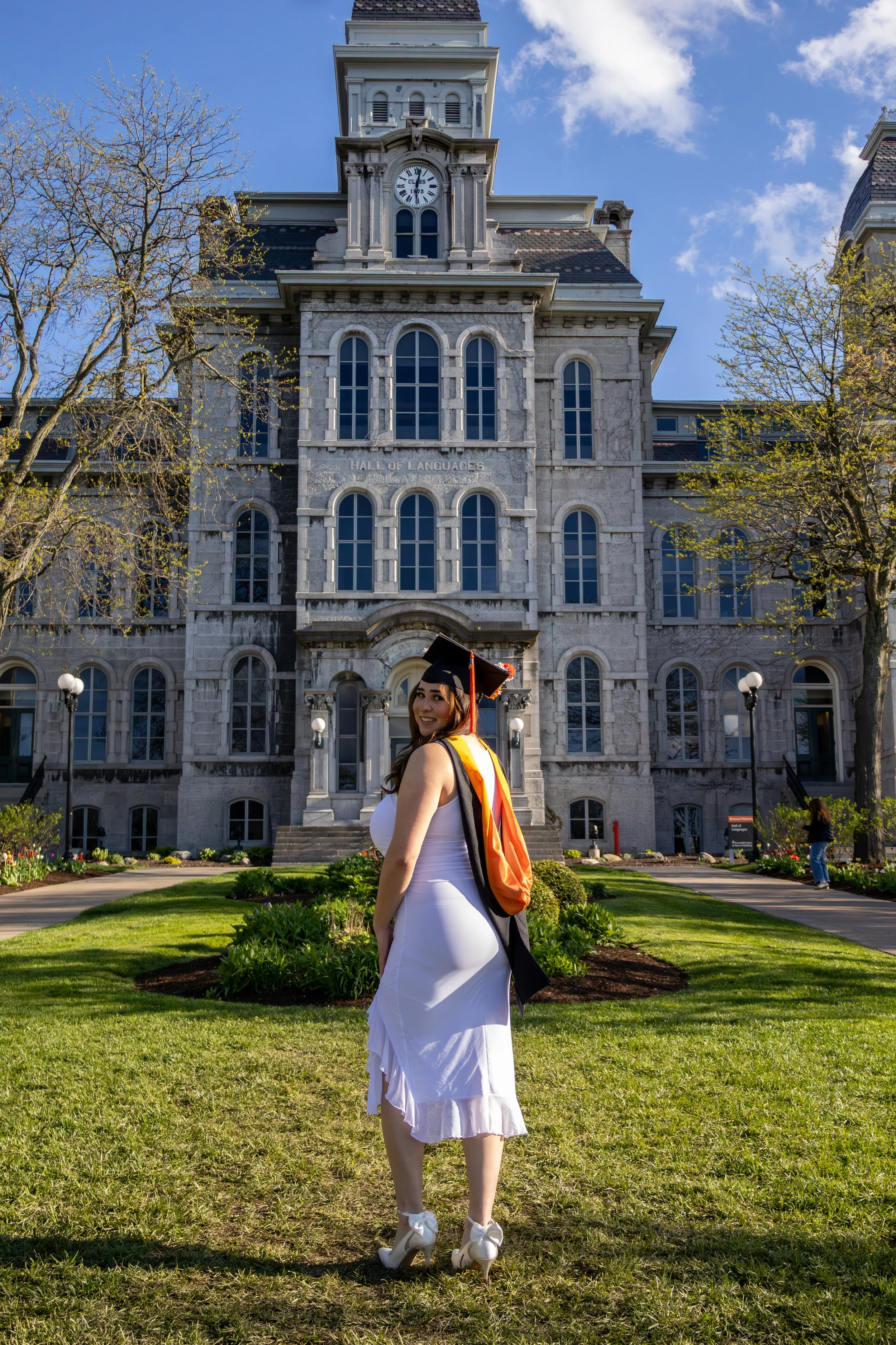 A woman in a white dress and high heels stands on a well-kept lawn in front of a large historic stone building with a clock tower, with trees and a blue sky in the background.