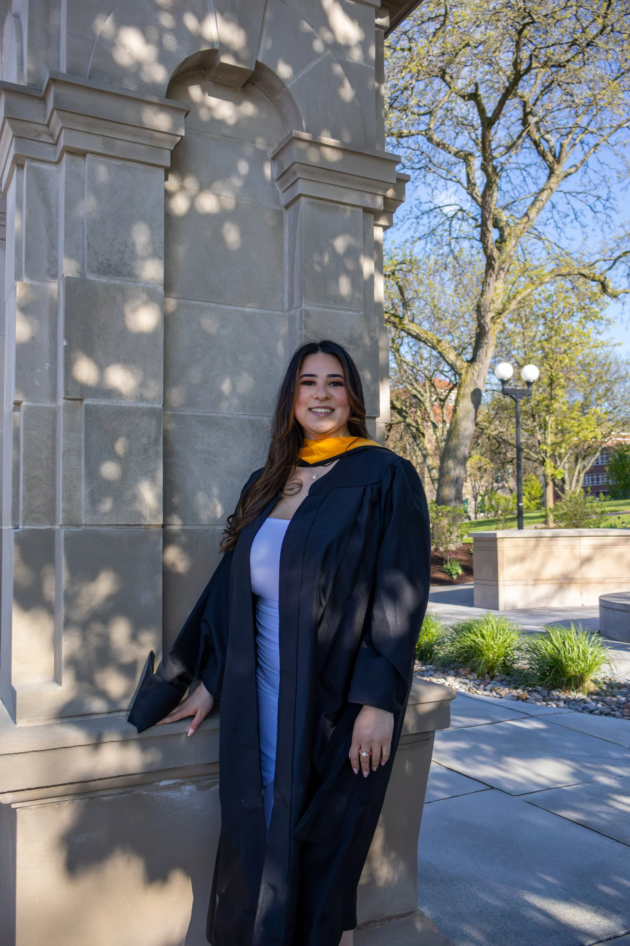 A young woman in a graduation cap and gown leaning against a stone building with trees and a lamppost in the background.