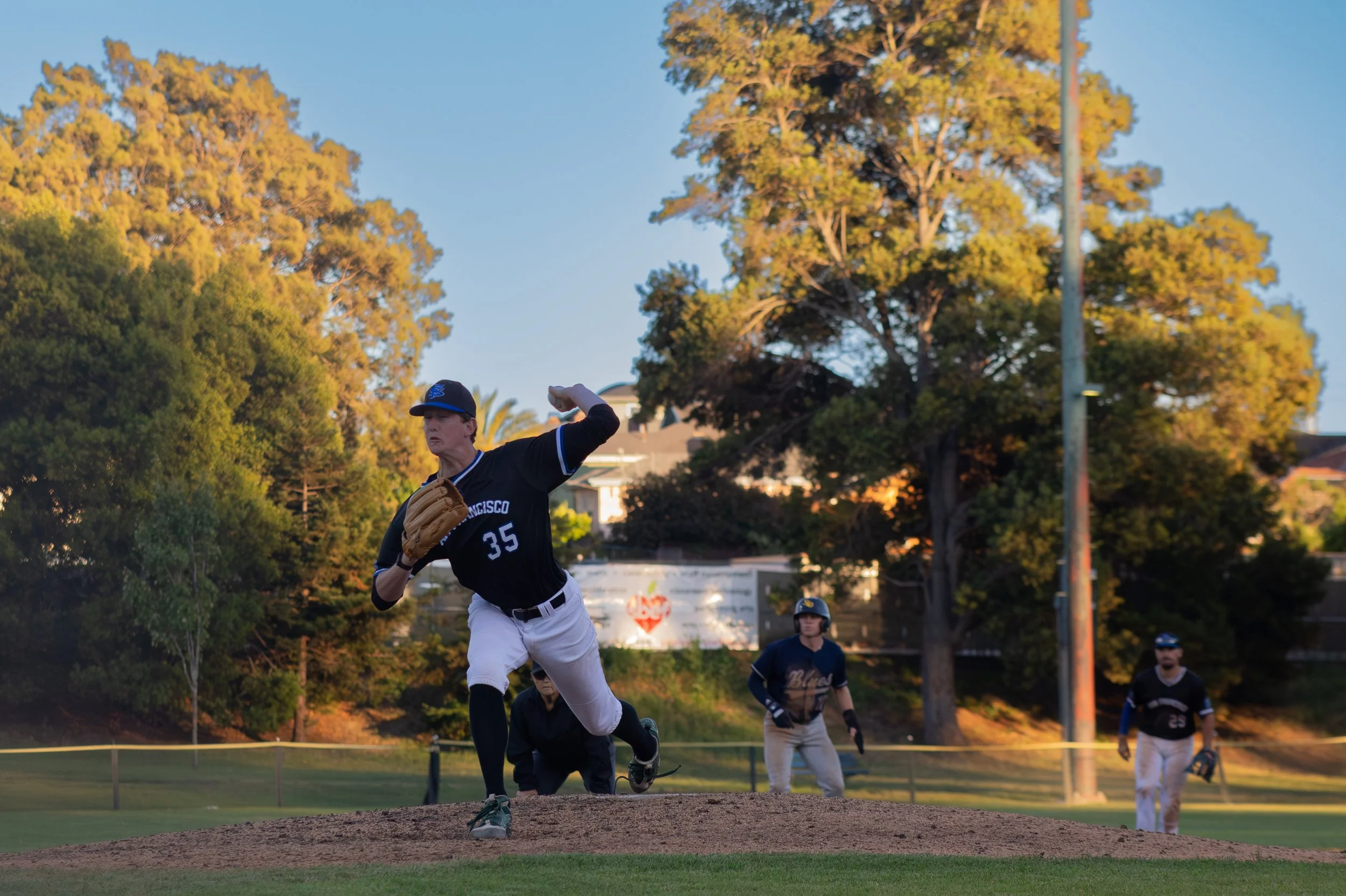 A baseball player in a black and white uniform with the number 35 on his jersey is in the middle of pitching on the mound. Other players in similar uniforms are visible in the background, along with trees and a clear sky.