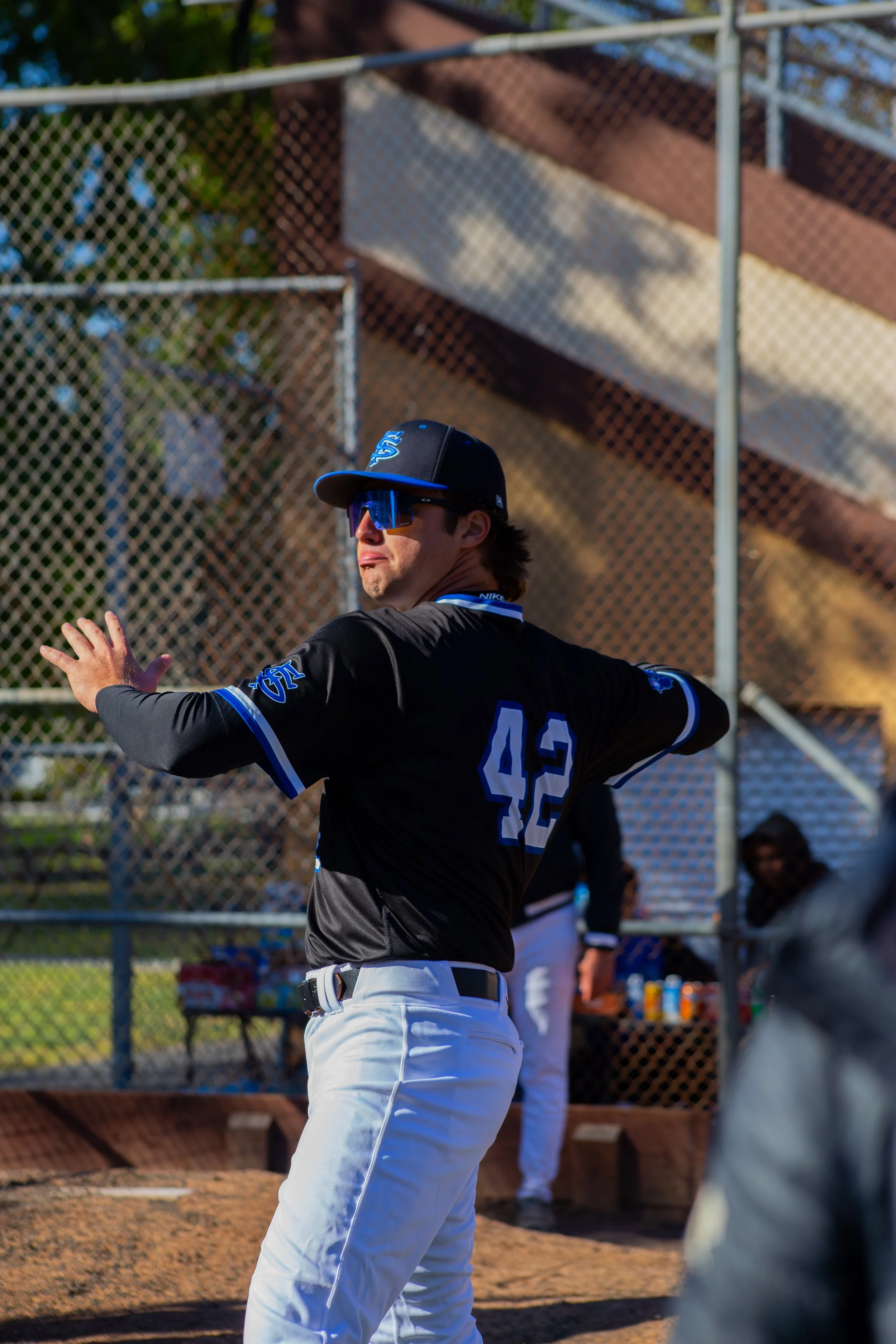 A baseball player wearing a black uniform with the number 42 and logo patches, a black cap with blue accents, and blue sunglasses, preparing to bat during a game at the baseball field.
