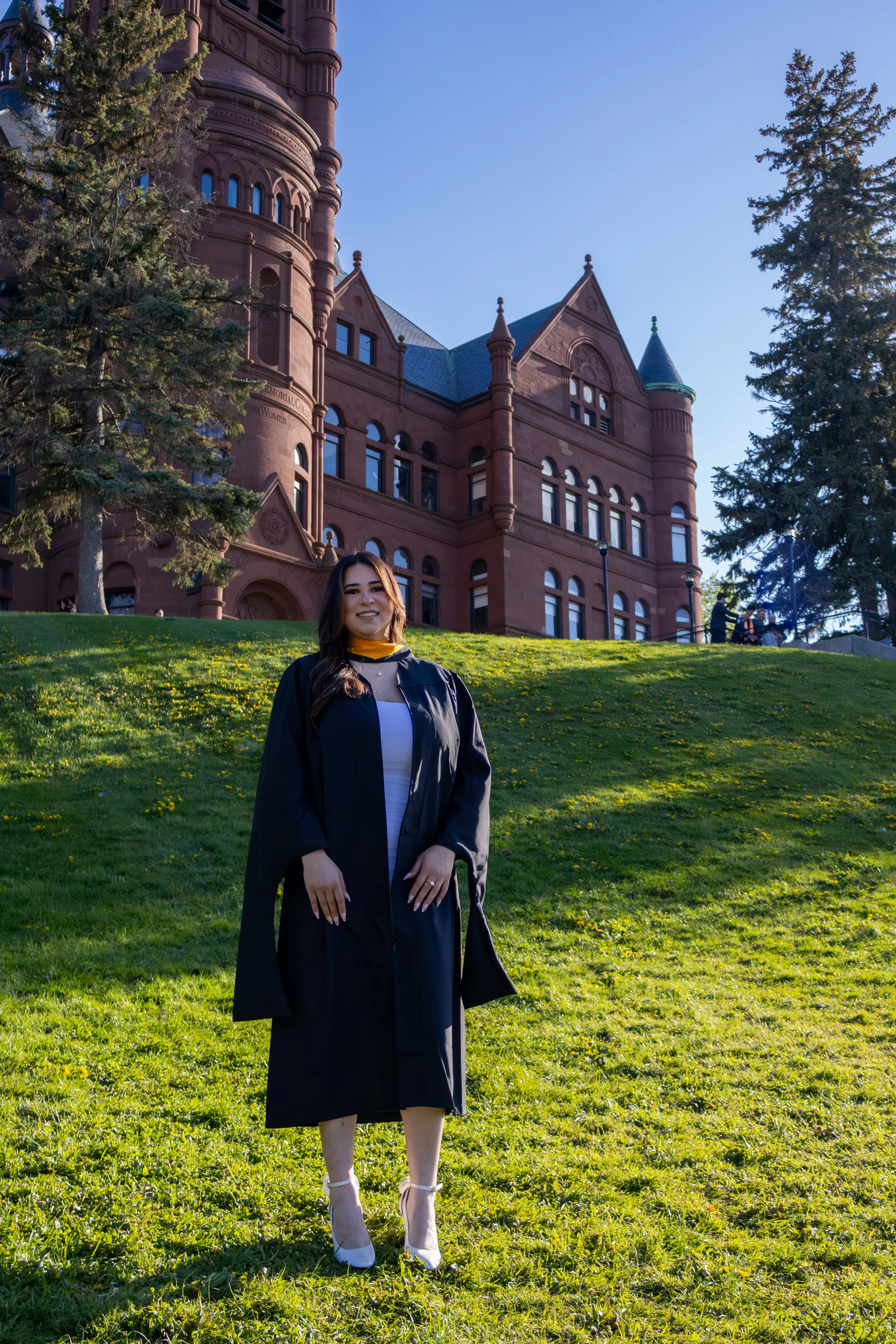 A young woman in a graduation cap and gown smiling outdoors on a grassy hill, with a historic red sandstone building and tall trees in the background.
