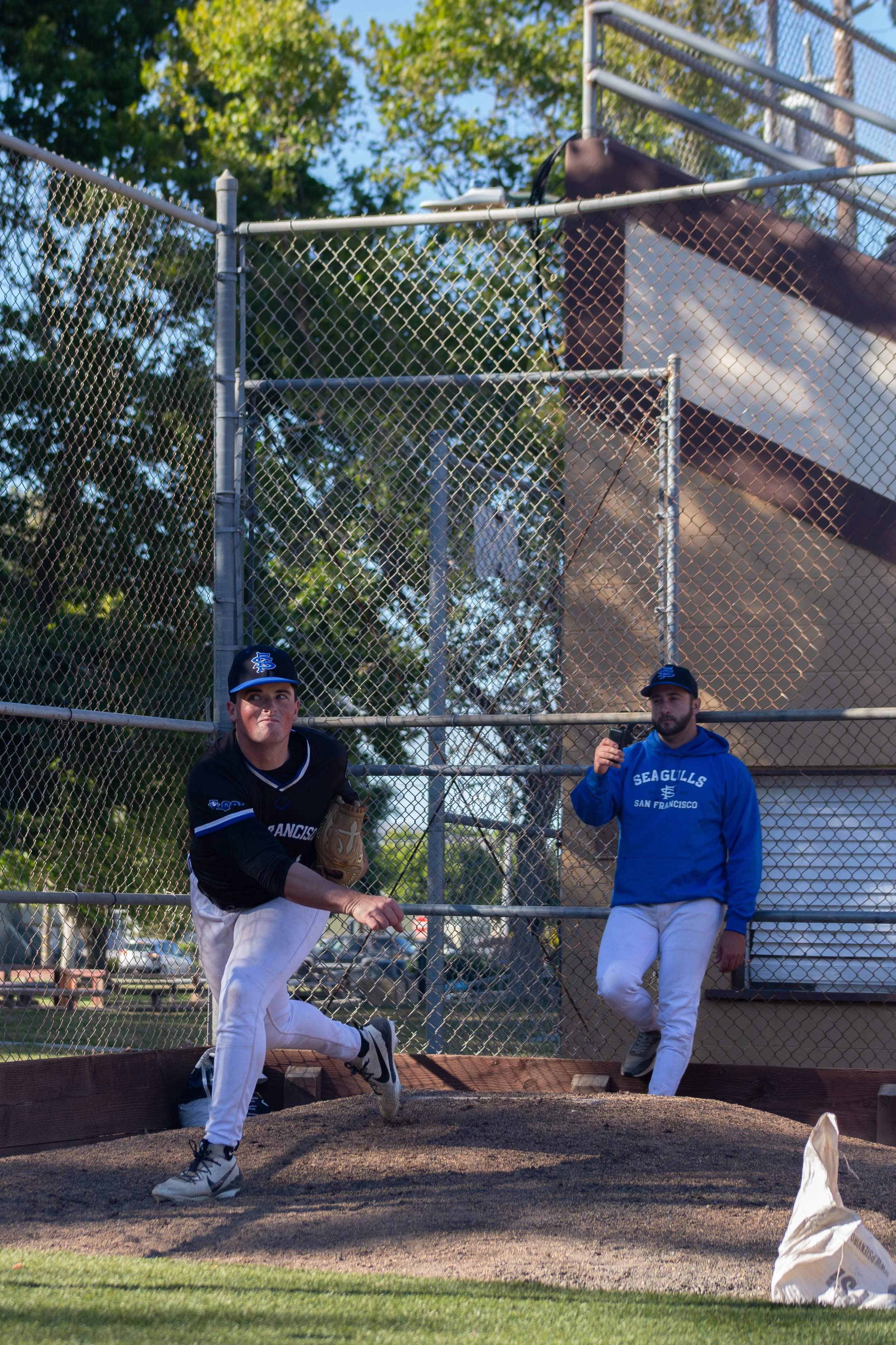 Two baseball players warming up before a game, one in black and one in blue, on a baseball field with a chain-link fence and trees in the background.