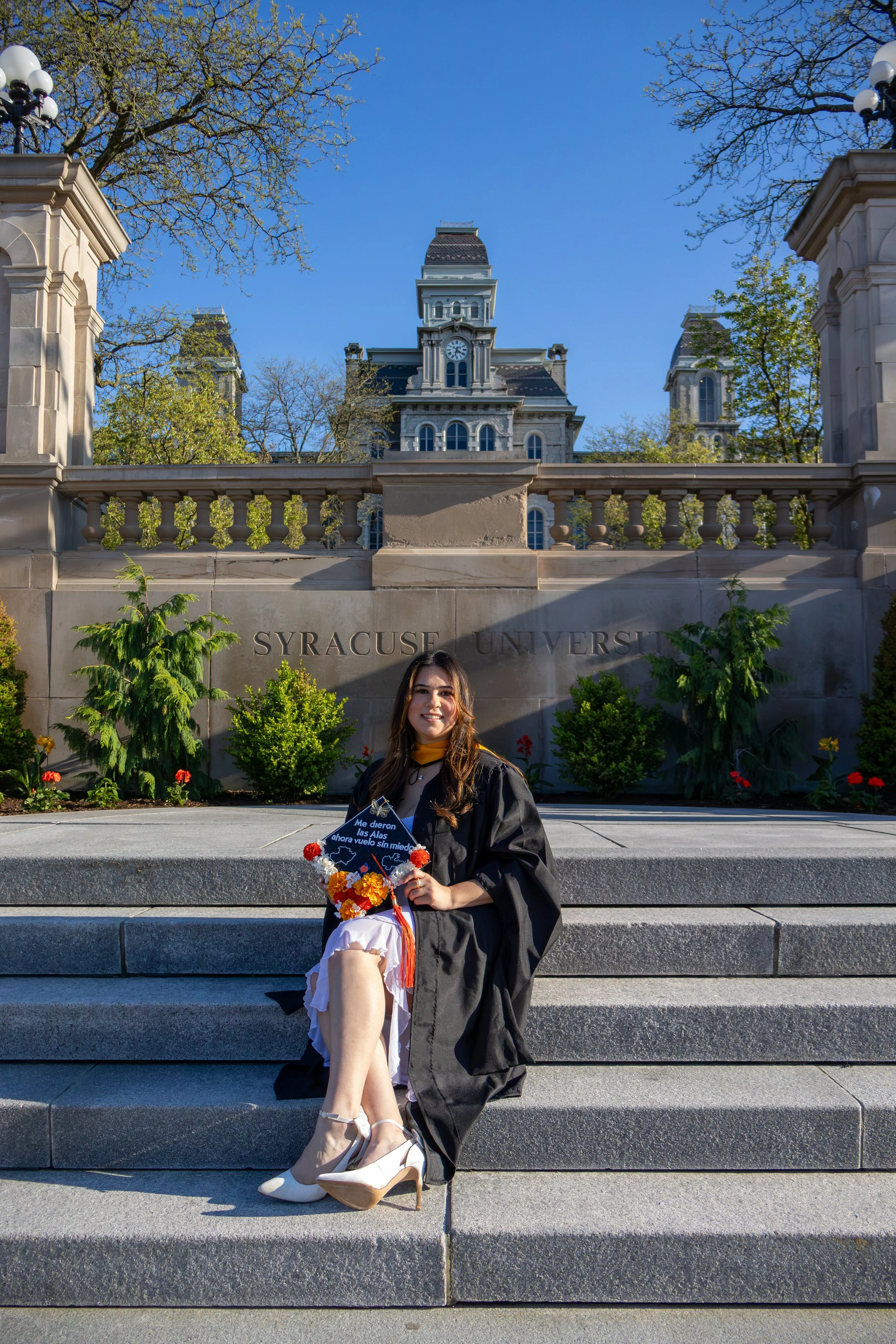 A young woman in a graduation cap and gown sitting on the steps of Syracuse University, holding a diploma and flowers, with a historic university building and trees in the background.