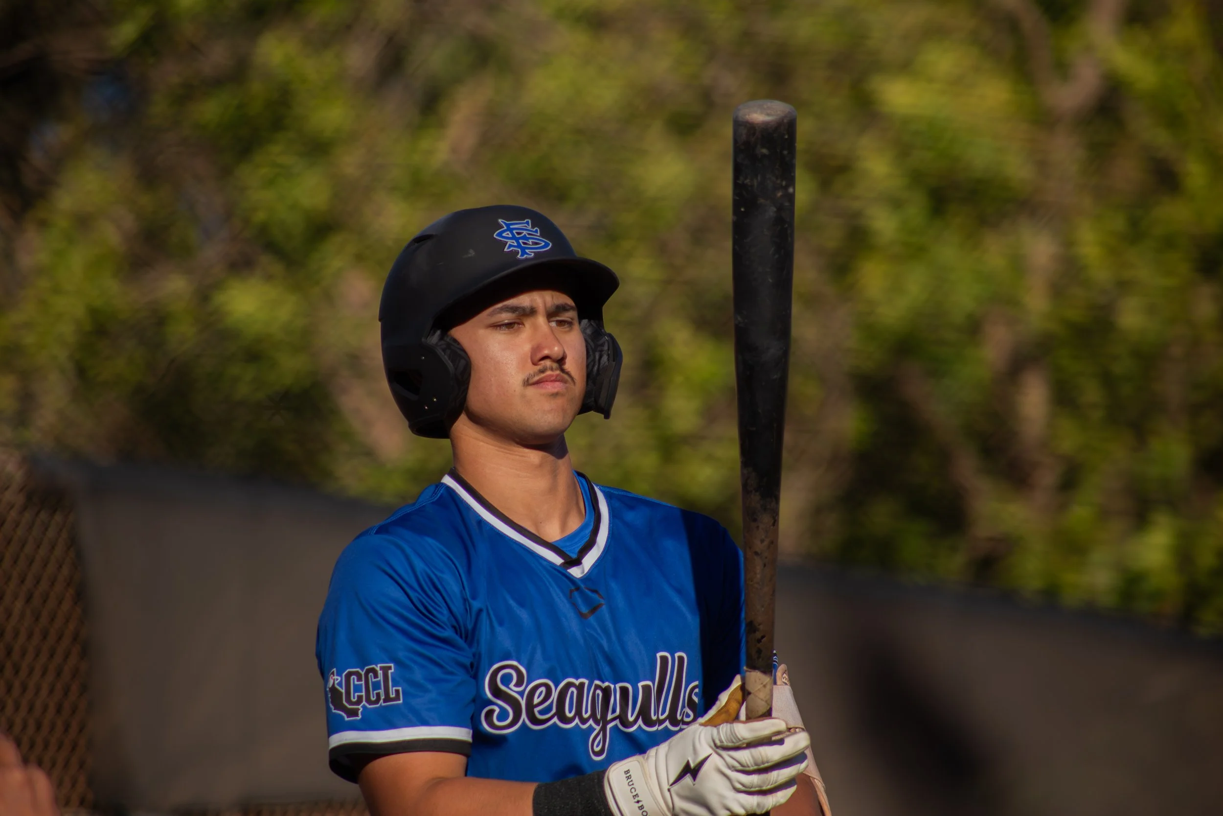 A young male baseball player in a blue uniform and batting helmet holding a baseball bat, standing outdoors with a blurred background of greenery.