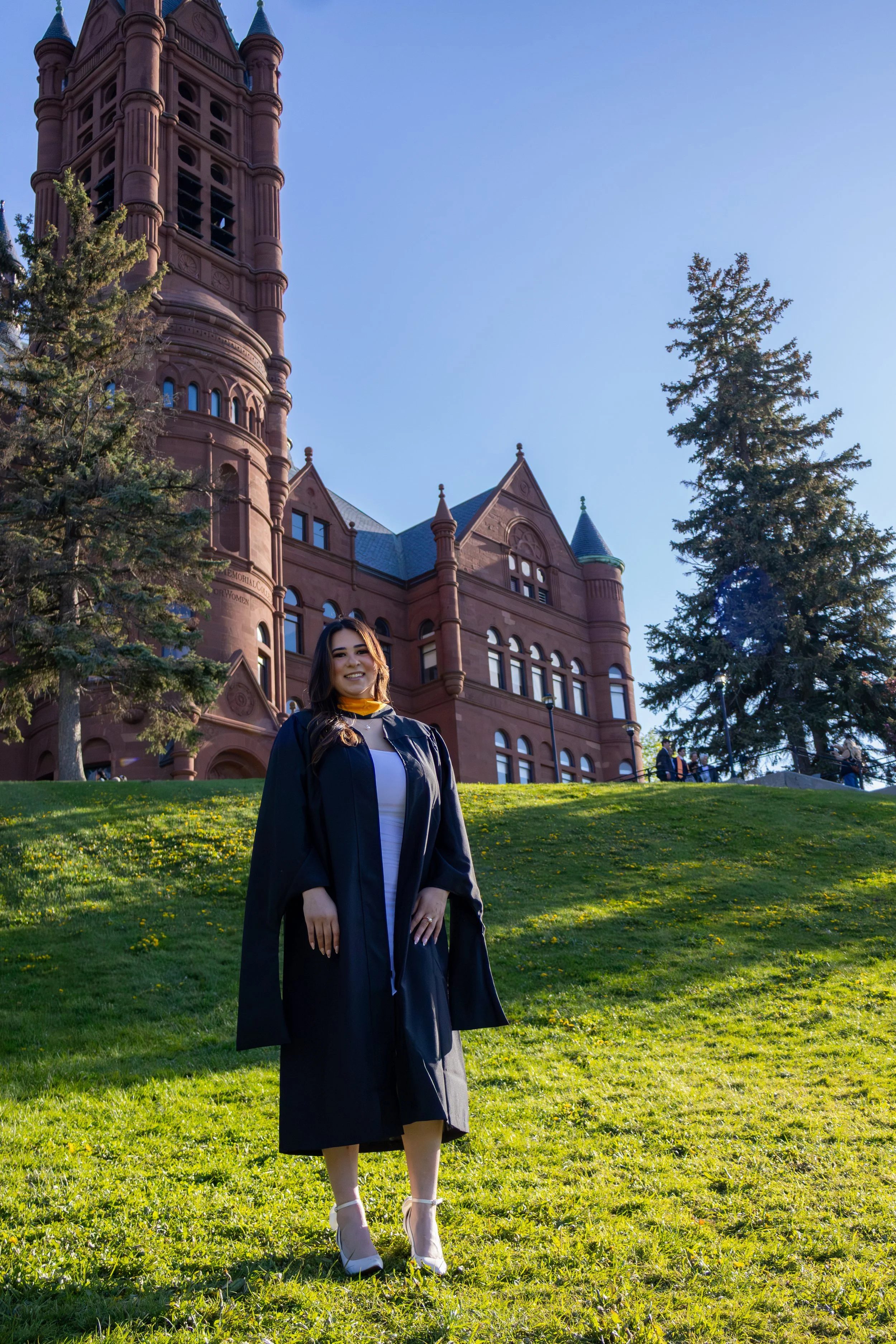 A young woman in a graduation gown and cap standing on a grassy area in front of a historic red brick university building with tall spires and windows, surrounded by trees and other people.
