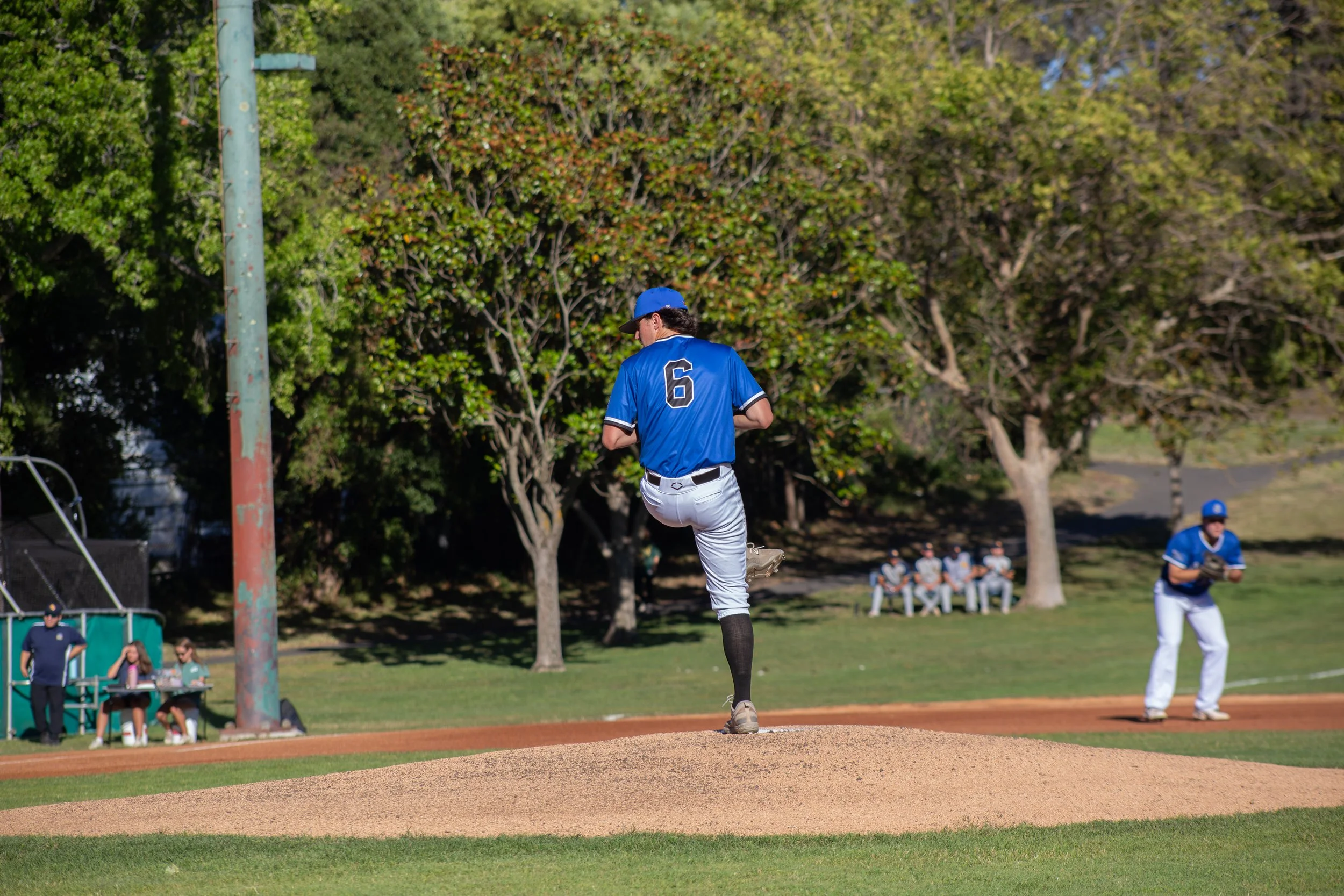 Baseball player in a blue jersey and white pants pitching from the mound with spectators and trees in the background.